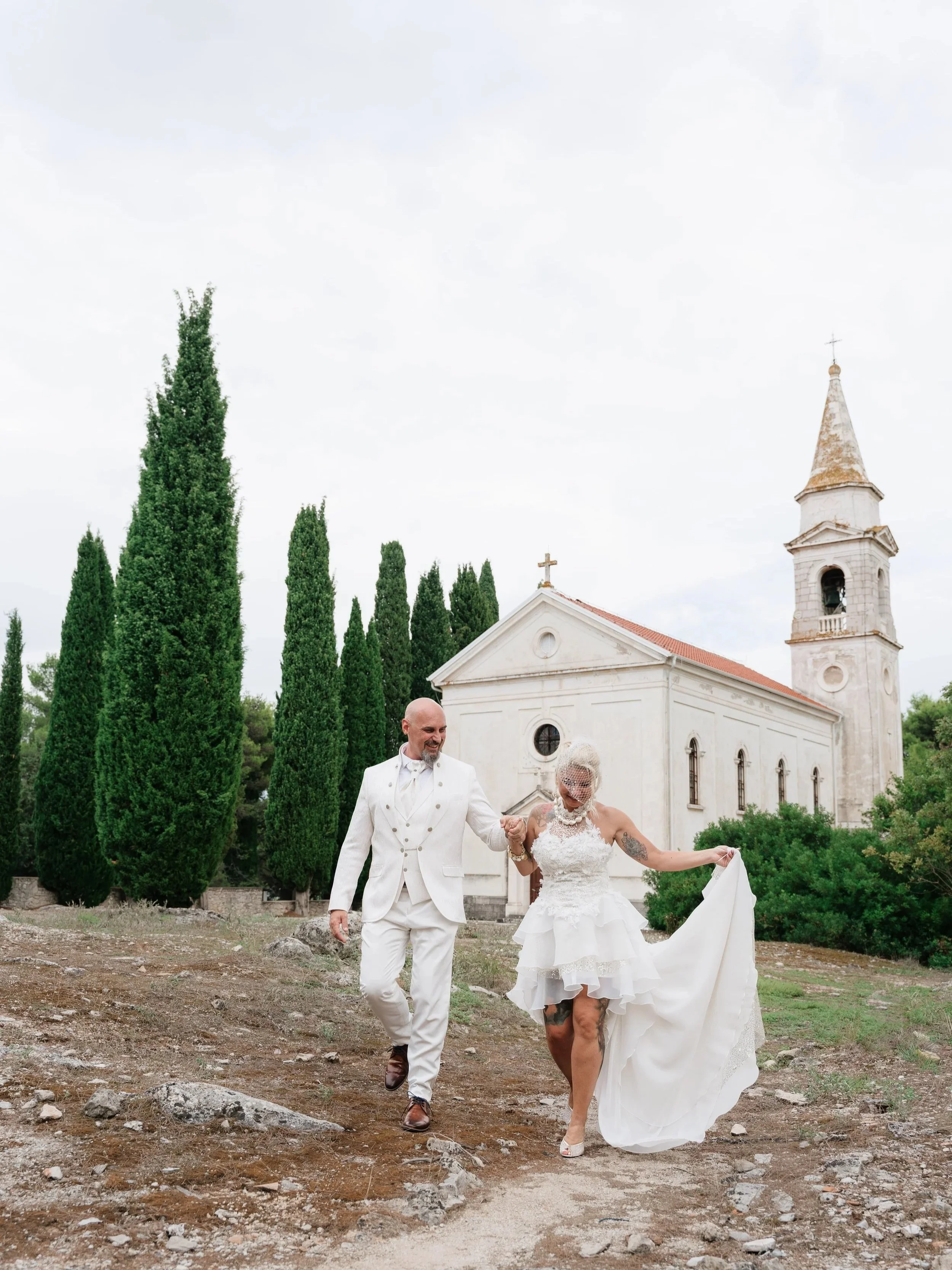 A newlywed couple walking outdoors in front of a church with tall cypress trees. The bride is wearing a white wedding gown and heels, holding the groom's hand, who is dressed in a white tuxedo with brown shoes. The bride is lifting her dress, reveali