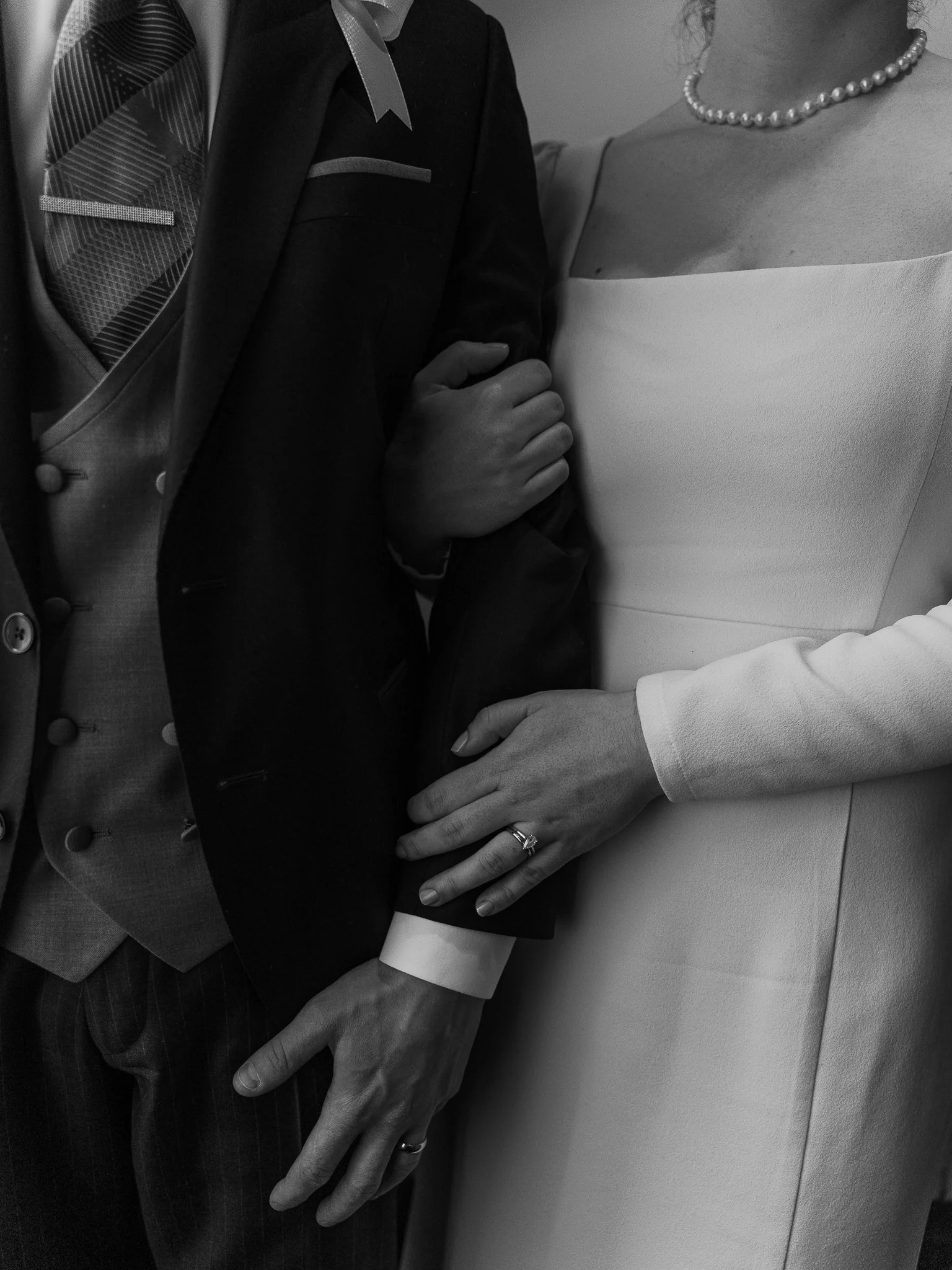 Close-up of a man and woman holding hands, with the woman's hand displaying rings, including a wedding band. The woman is wearing a pearl necklace, and the man is dressed in a suit with a patterned tie and vest.