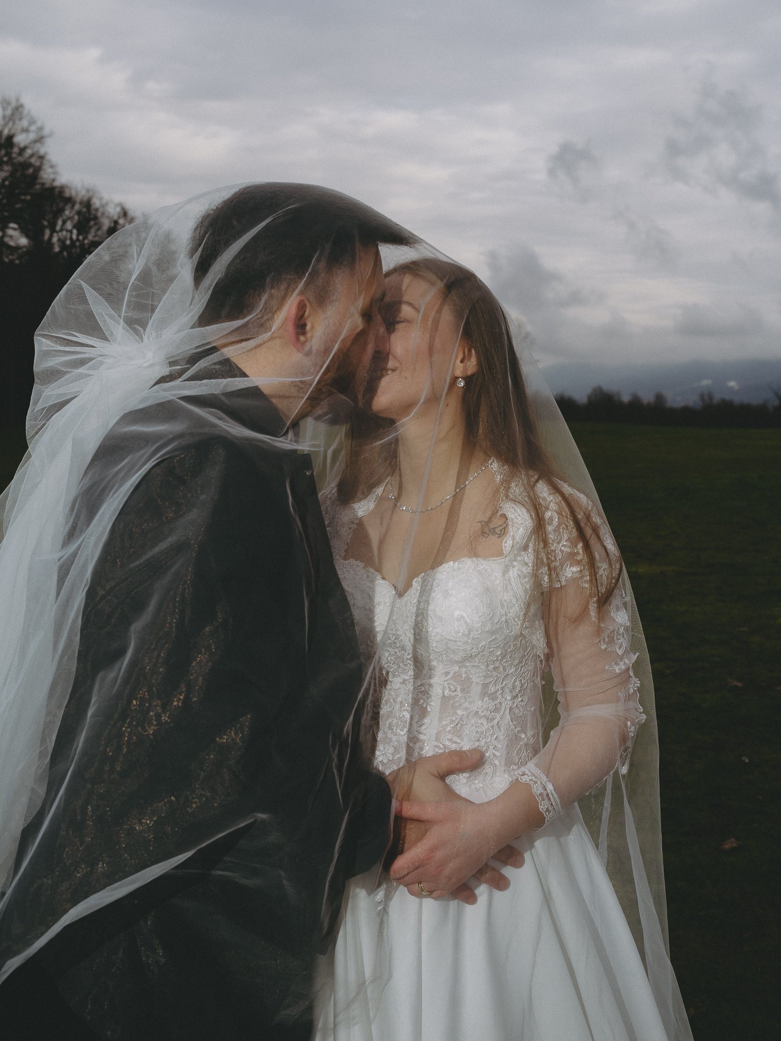 A couple dressed in wedding attire, under a veil, sharing an intimate moment outdoors on a cloudy day.