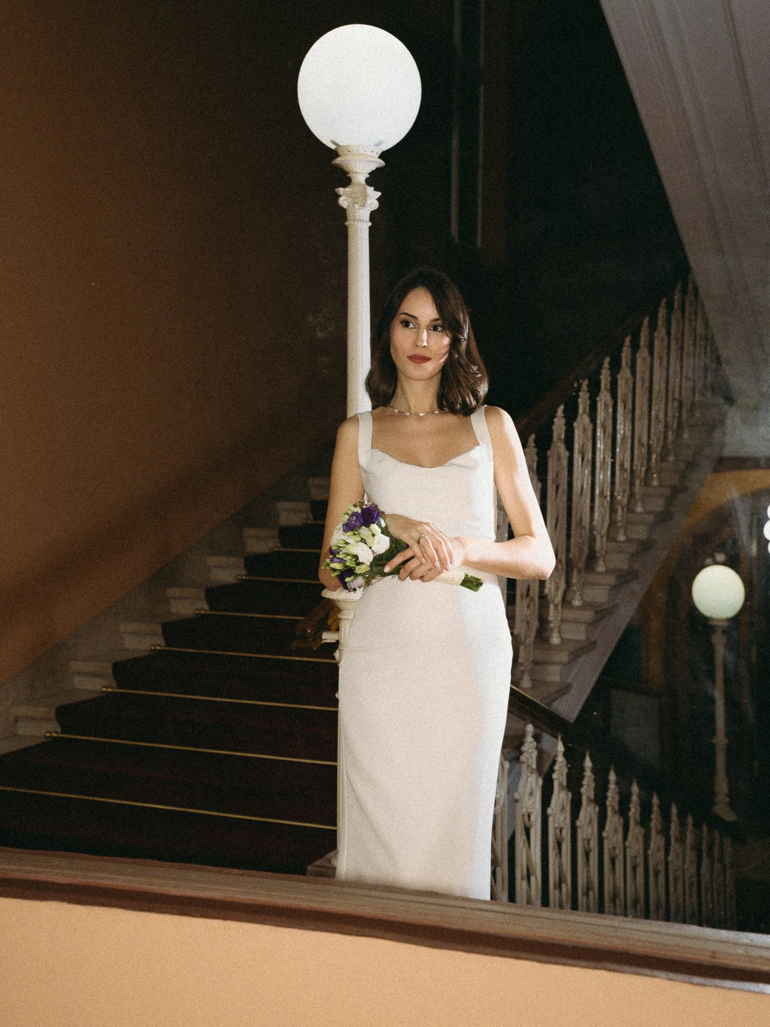 Woman in a white dress holding a bouquet of purple and white flowers, standing on a staircase in an indoor setting with vintage lighting.