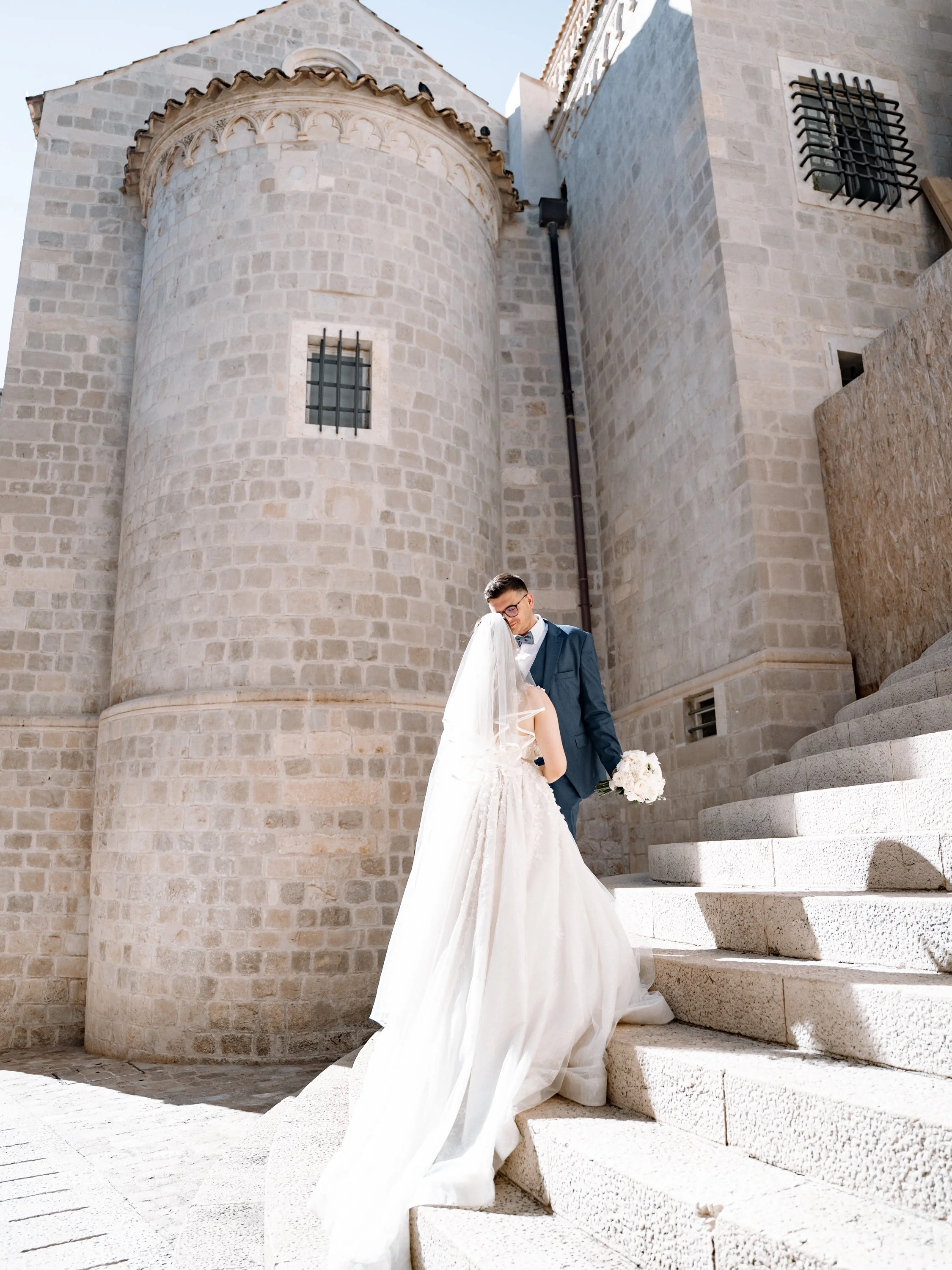 A bride and groom sharing a moment on stone stairs in front of a historic stone building, possibly a church, with large rounded towers and barred windows, during the daytime.