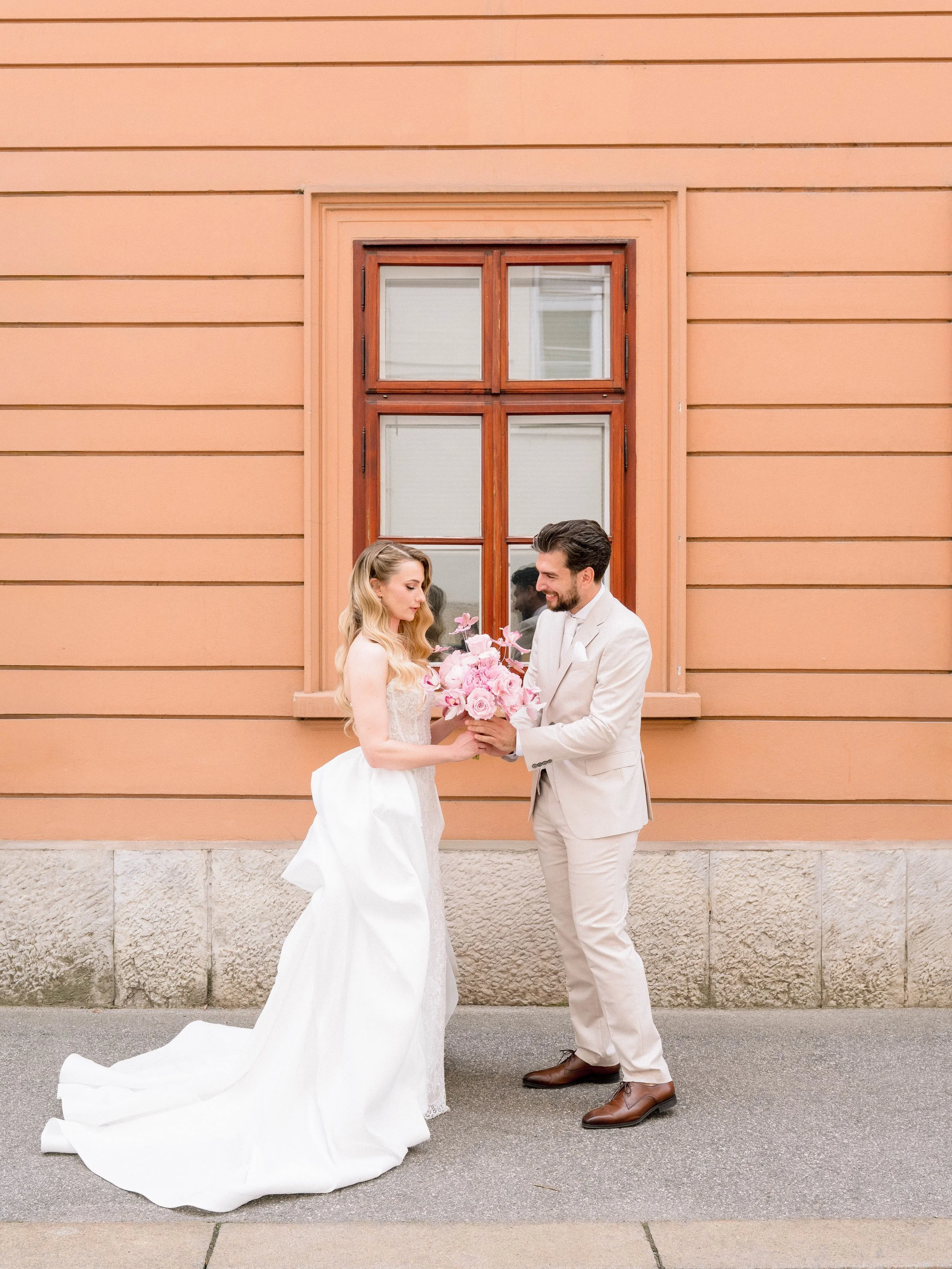 A bride and groom standing outside a peach-colored building with a window, exchanging flowers, with cobblestone and sidewalk underneath.