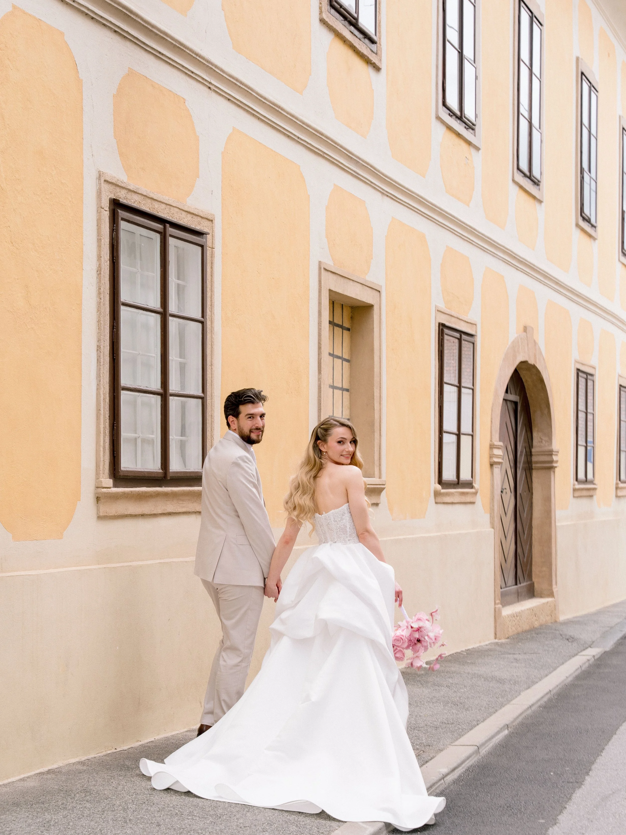 A happy couple dressed in wedding attire holding hands and walking outside the historic European-style building, with smiling expressions and the bride holding a bouquet of pink flowers.
