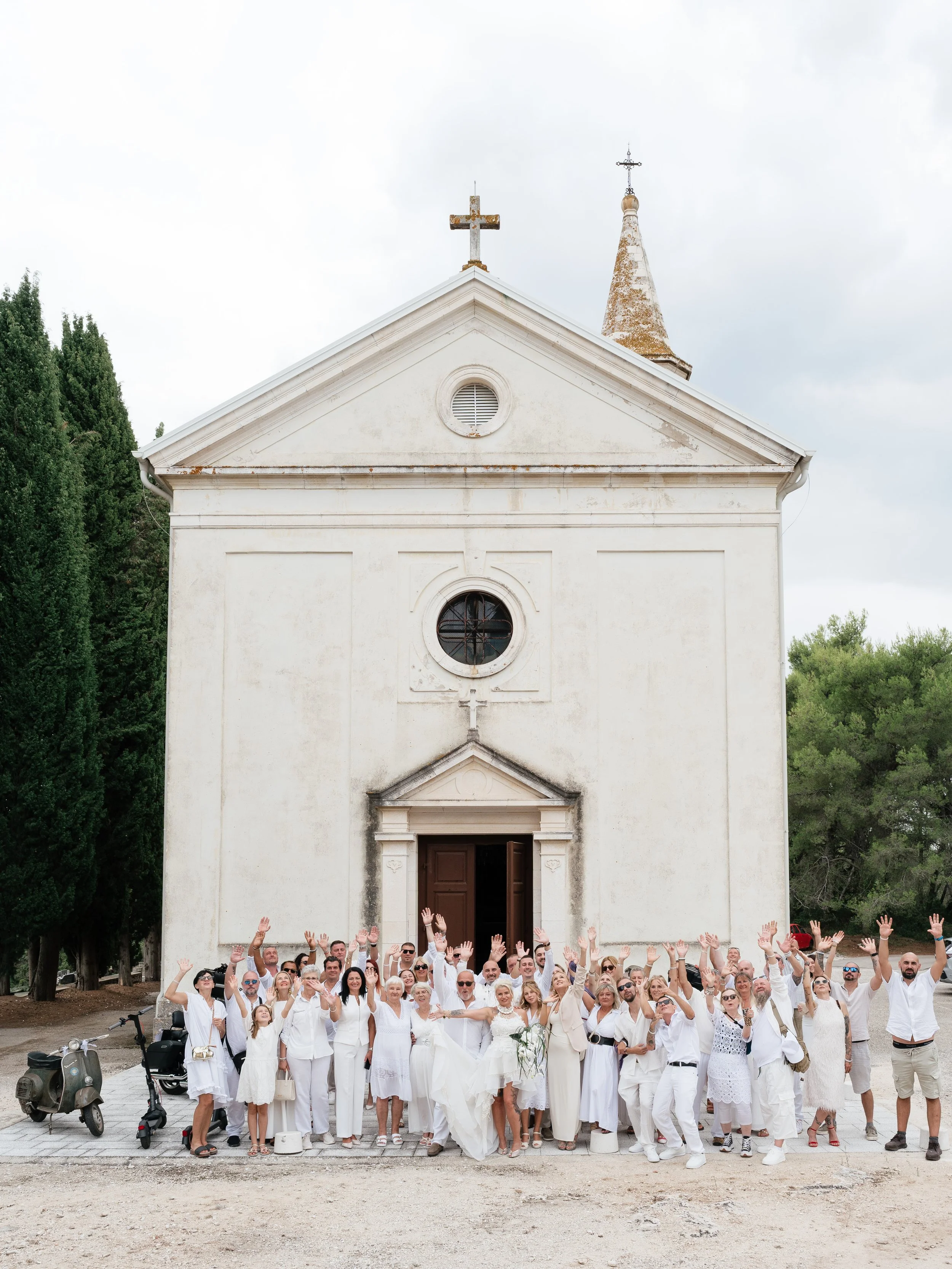 A large group of people dressed in white are gathered in front of a small church, raising their hands in celebration or greeting.