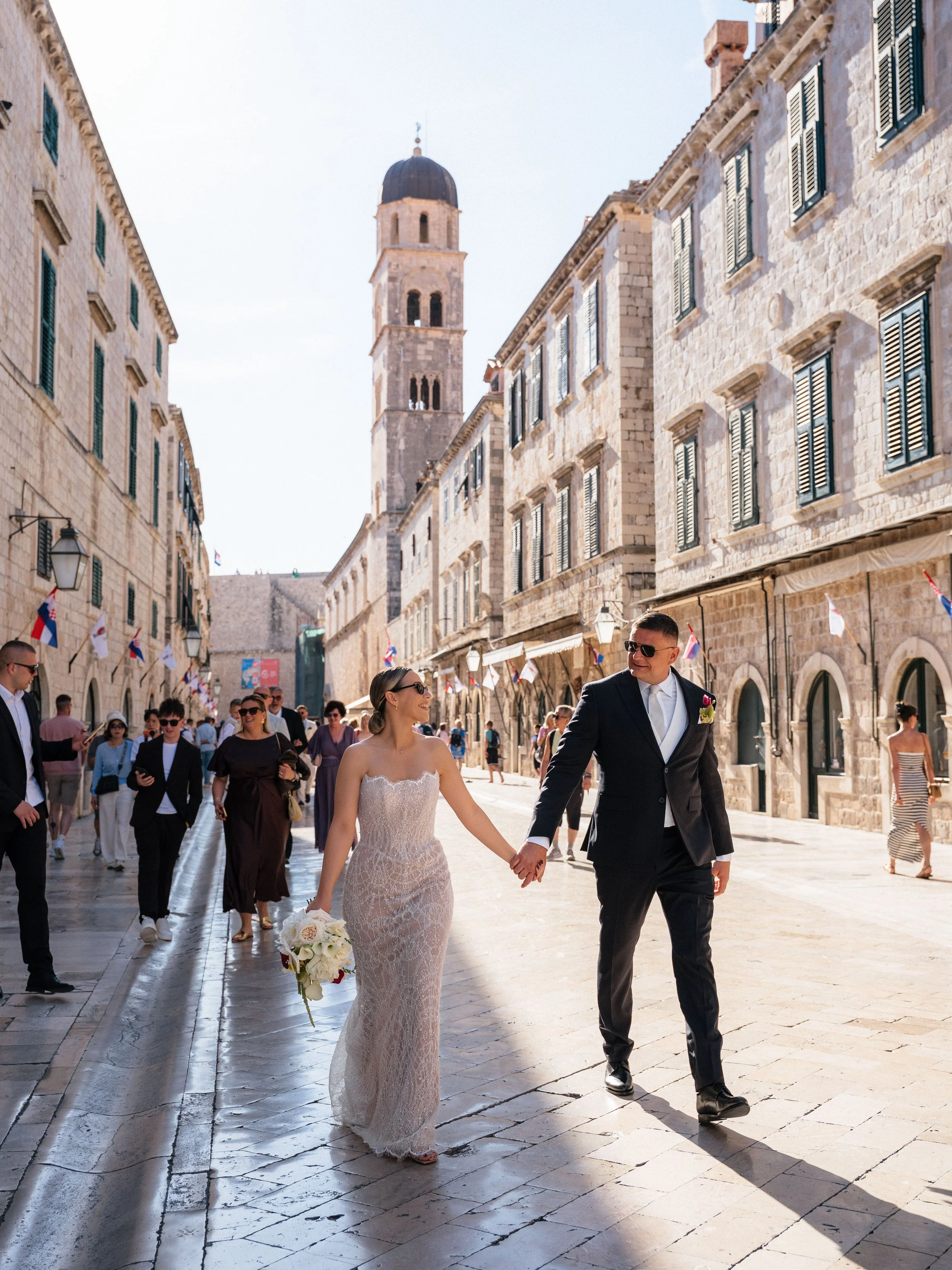 Couple in wedding attire walking hand-in-hand on a sunny stone street Stradun (in Dubrovnik, Croatia) surrounded by people, historic stone buildings, flags, and a church tower in the background.