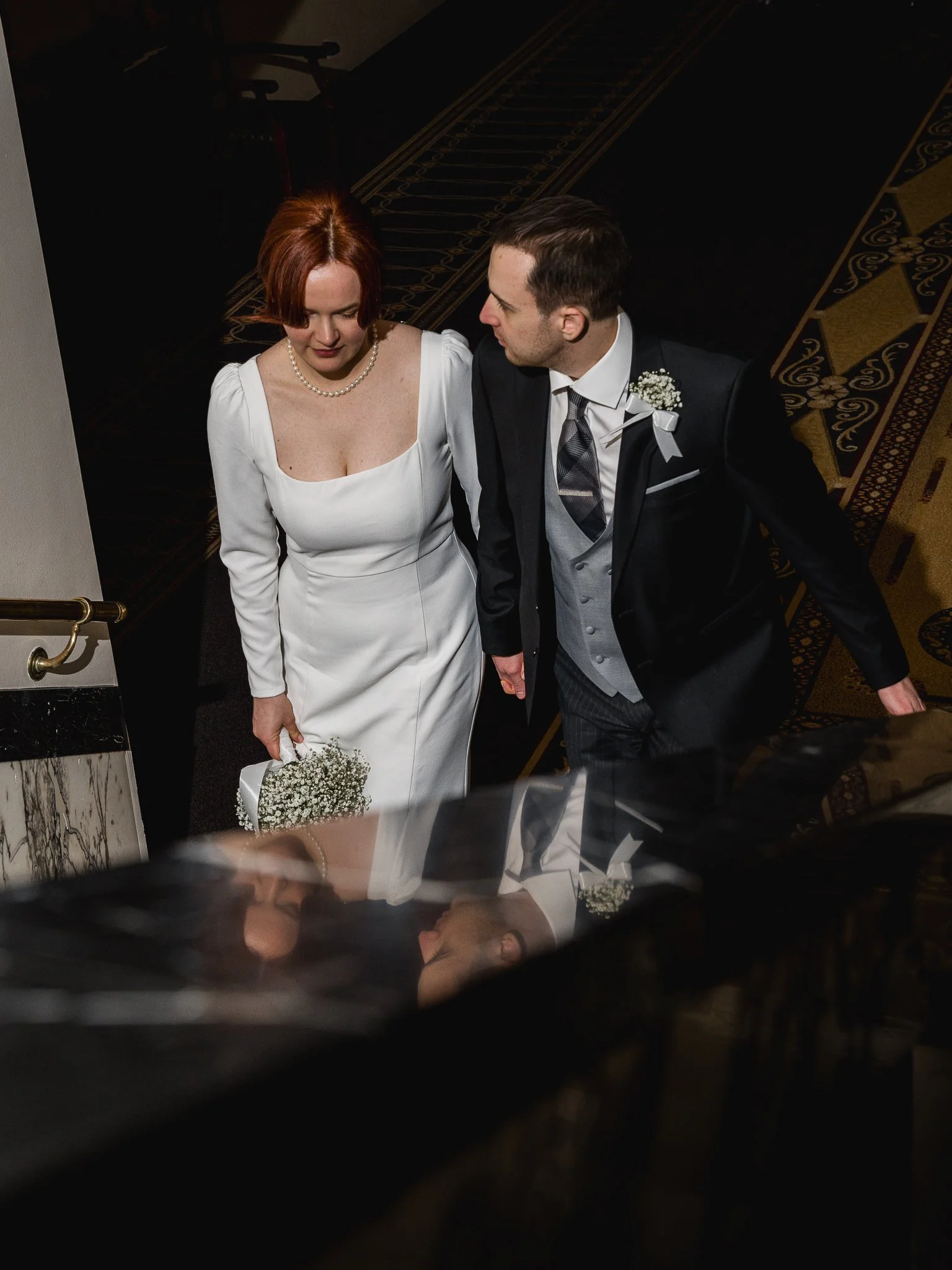 A bride and groom walk up a staircase during their wedding ceremony. The bride is holding a small bouquet of white flowers and is wearing a white wedding dress with long sleeves, pearl necklace, and red hair. The groom is dressed in a black suit with