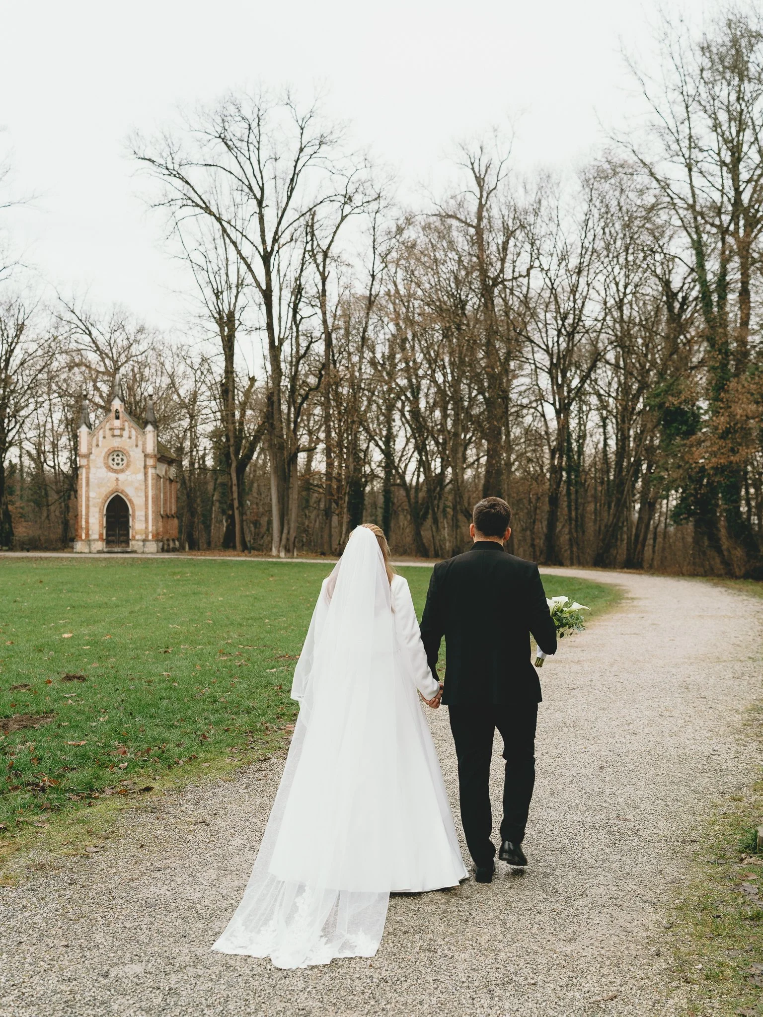 Bride and groom walking hand in hand on a gravel path near trees and a small chapel on an overcast day.