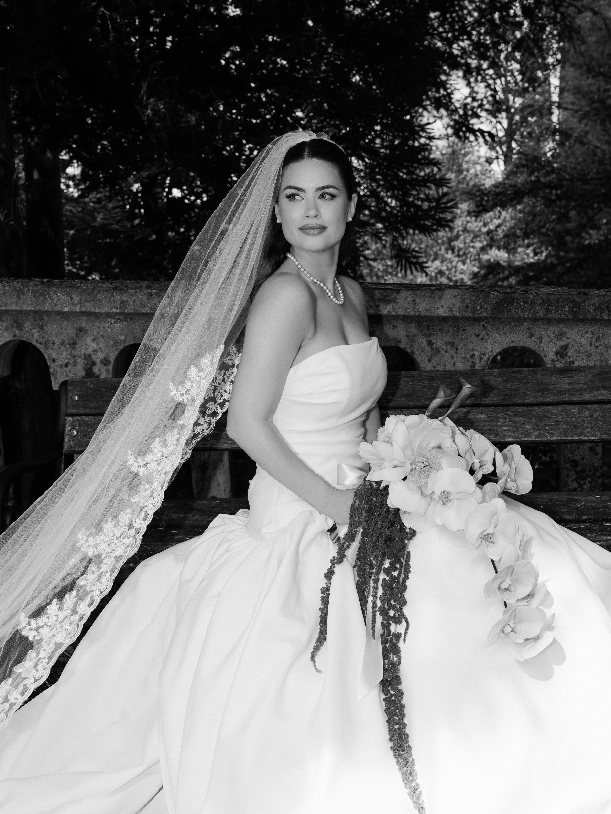 Black and white photo of a woman in a wedding dress with a veil, sitting on a bench outdoors, holding a bouquet, with trees and sky in the background.