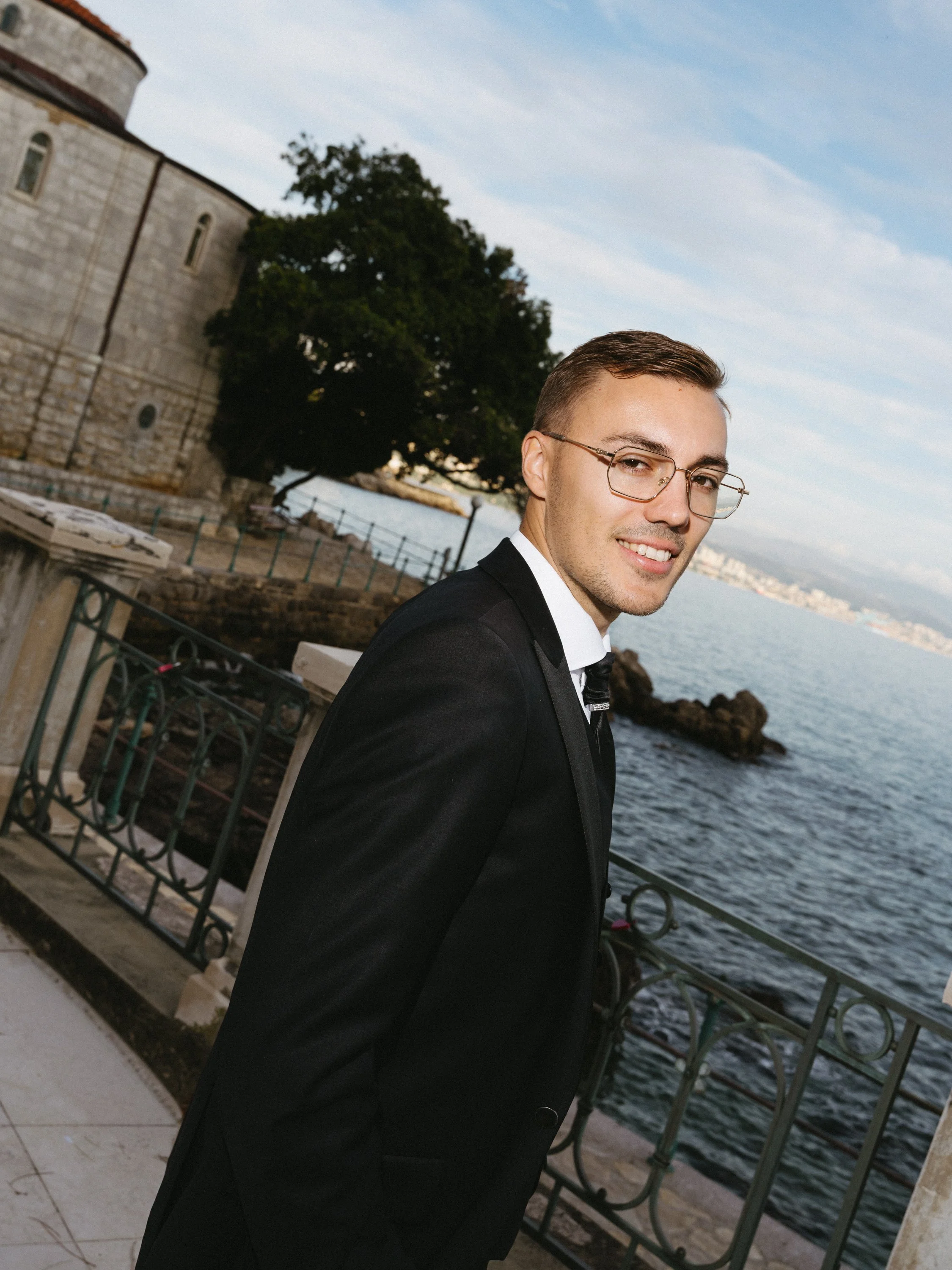 A young man in a black suit and glasses is standing by a waterfront with a castle and large tree in the background.