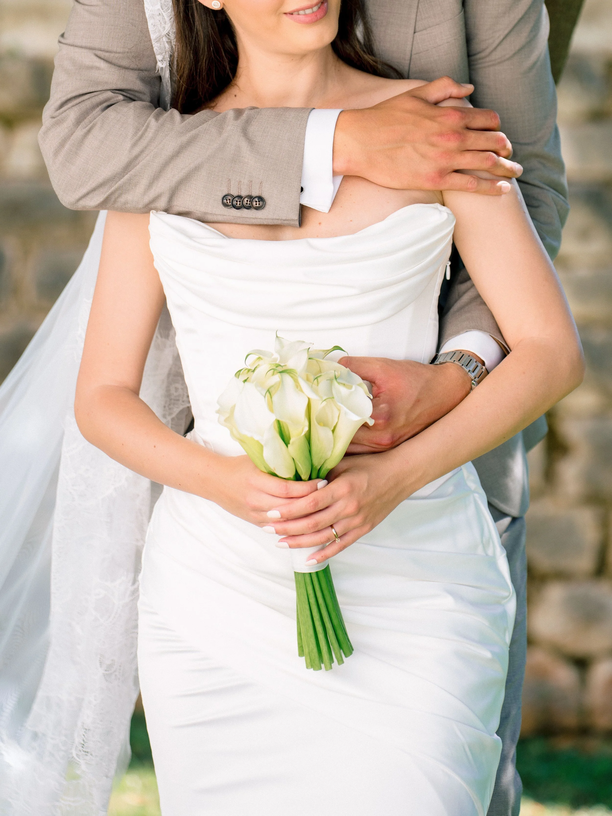 A bride in a white wedding dress holding a bouquet of calla lilies, standing next to a man in a beige suit, with his arm around her.