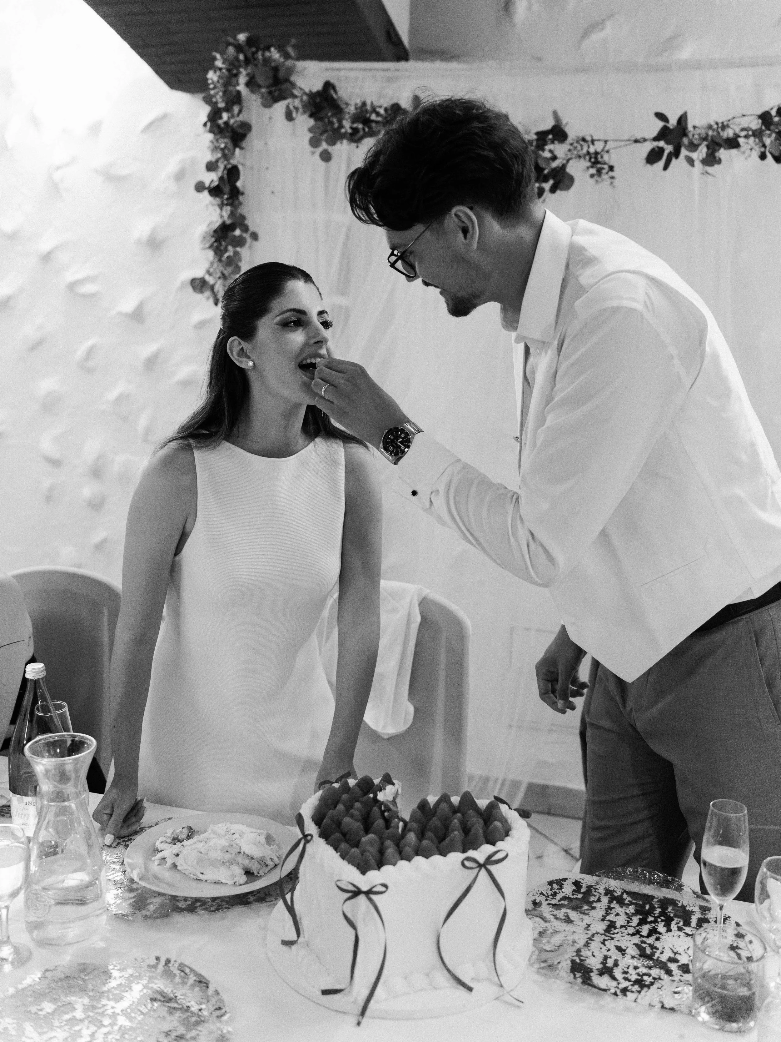 A man in glasses and a white shirt feeding a woman in a white dress at a celebration table. The table has a large cake with fruit and a box of assorted sweets, along with glasses of water and wine. Decor inside the room includes a floral garland.