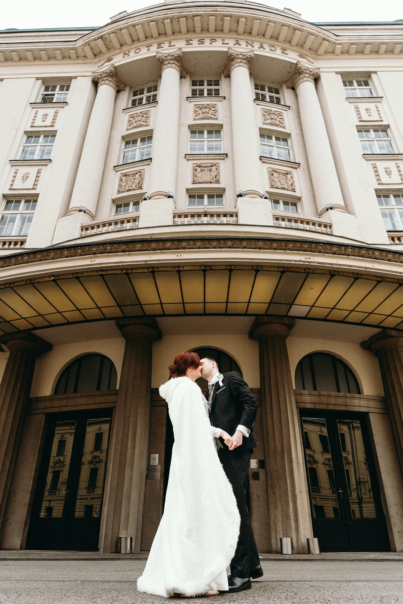 A bride in a white fur coat and a groom in a black suit sharing a kiss in front of a grand hotel building.