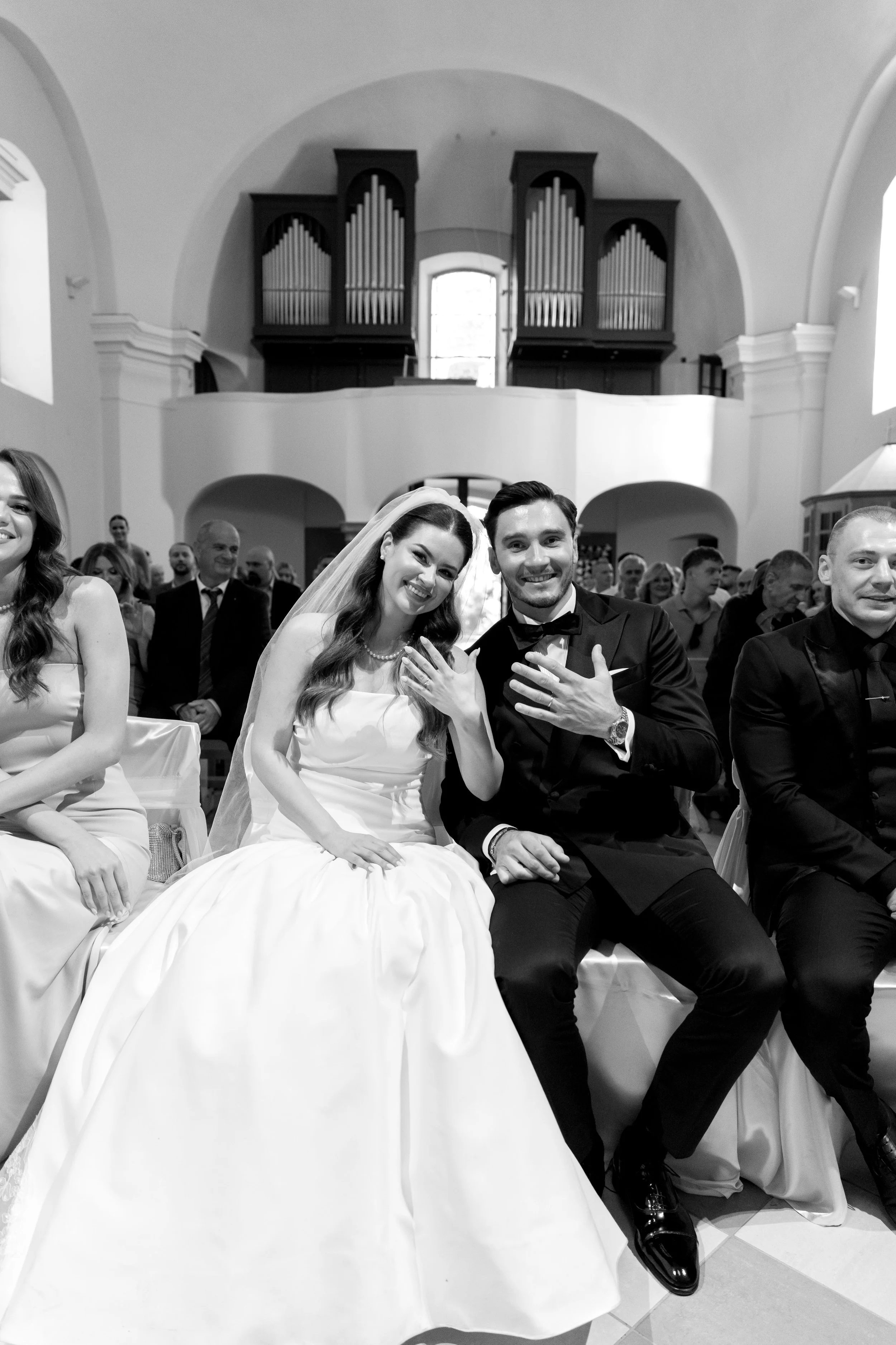 Black and white photo of a wedding ceremony with a bride and groom sitting together, surrounded by guests in a church. The bride is wearing a white wedding dress and veil, and the groom is in a black tuxedo. The background features an organ and arche