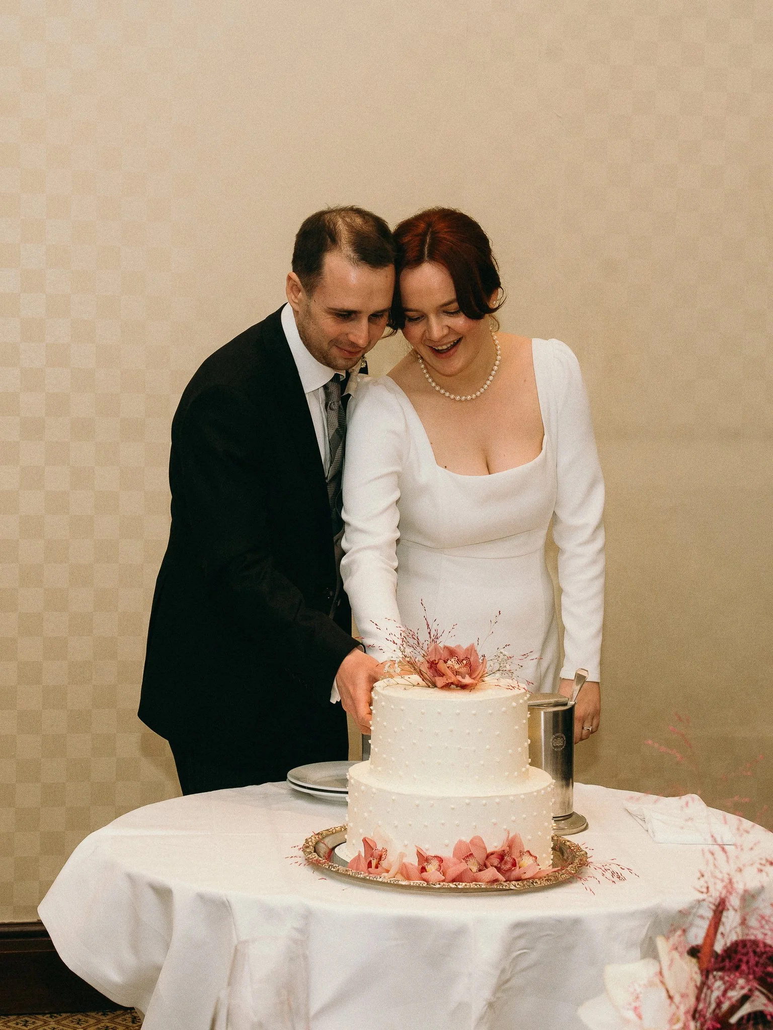 A bride and groom cutting their wedding cake together, surrounded by wedding decorations, with a two-tier white cake decorated with pink flowers and floral accents.