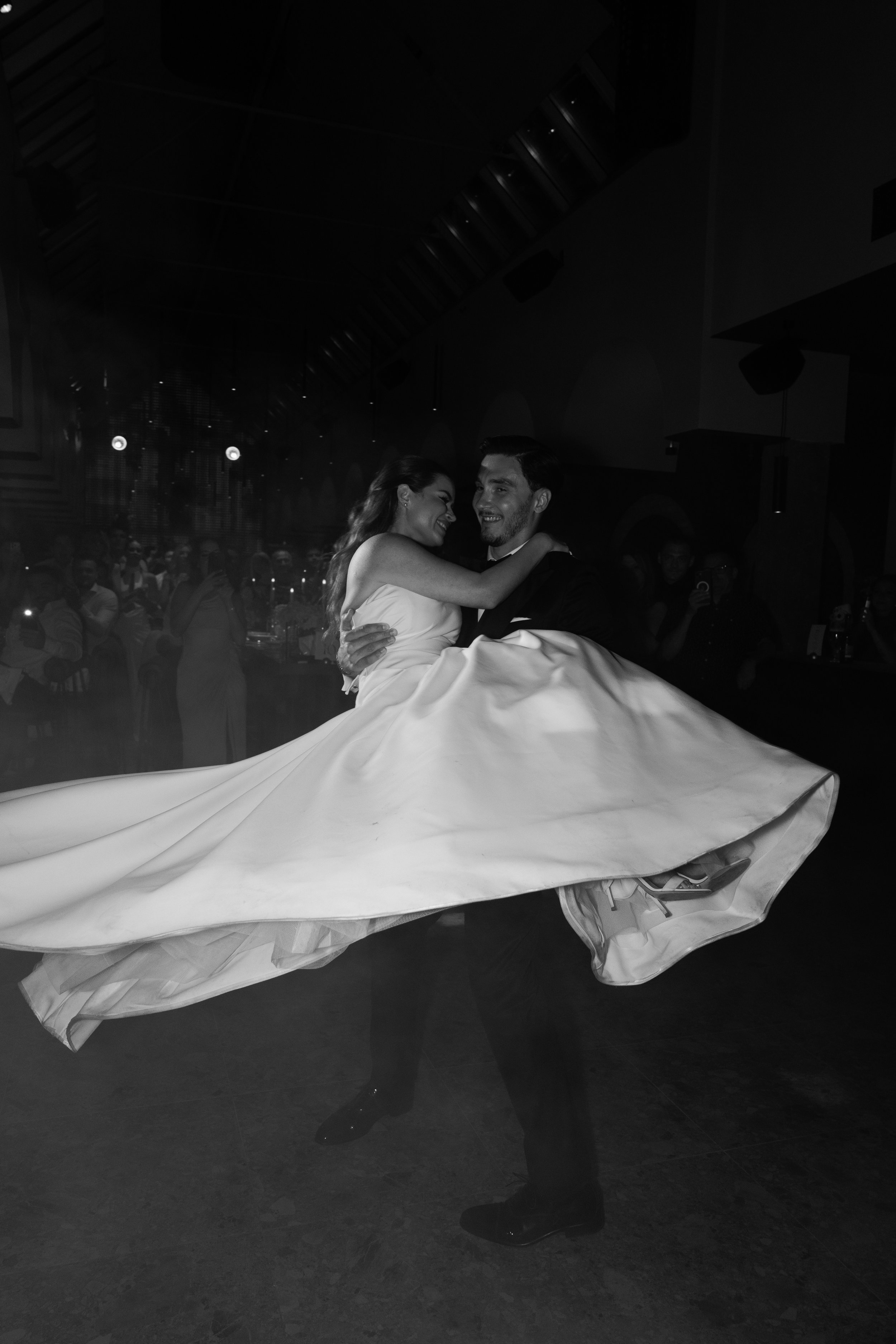A black and white photo of a man and woman dancing at a formal event. The woman is wearing a wedding dress with a long train, and the man is in a tuxedo. They are smiling and looking at each other, surrounded by an audience of watching guests in a di