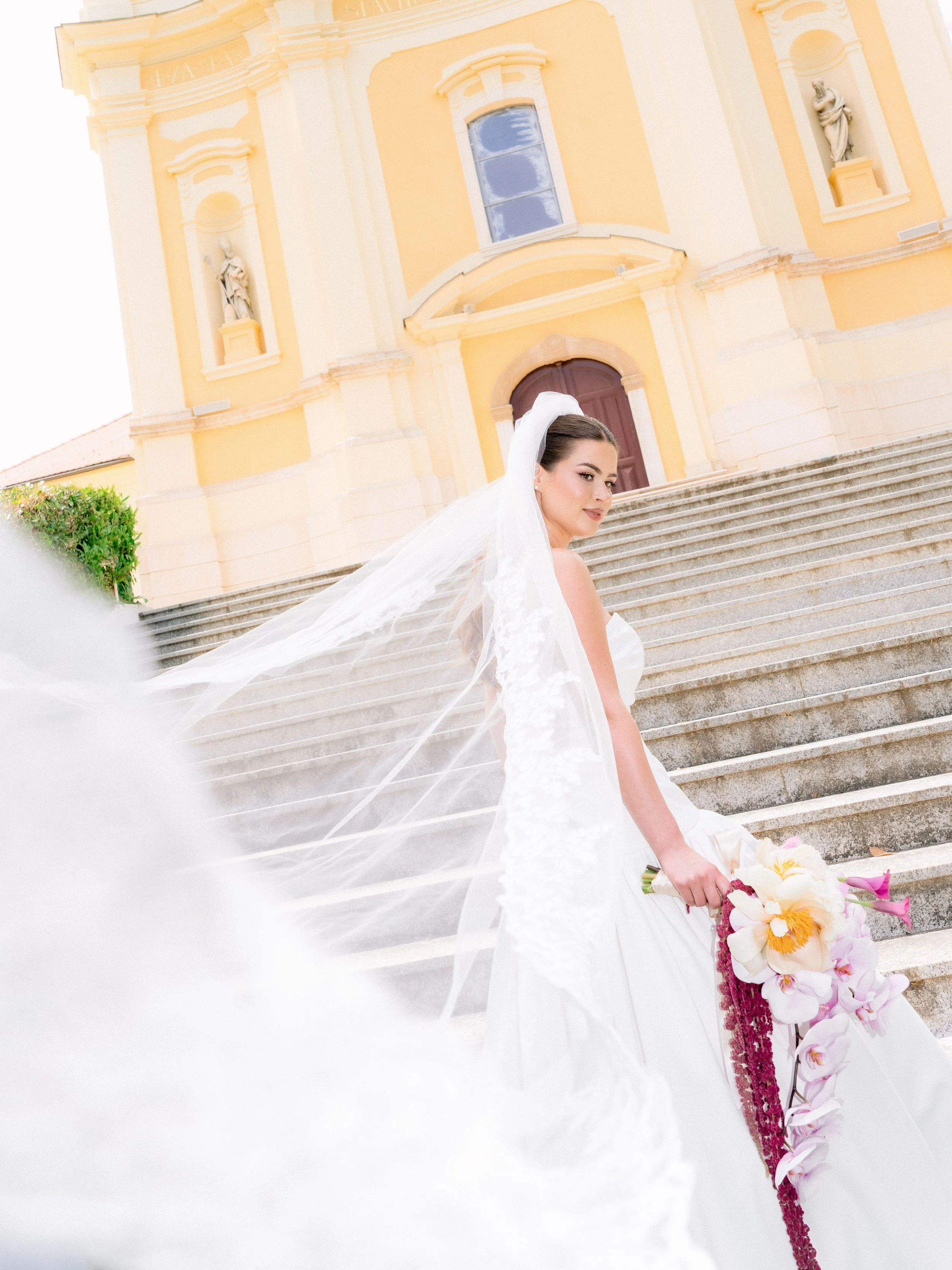 A bride in a white wedding dress and veil holding a floral bouquet stands on the steps of a yellow church with statues in niches, and steps leading up to the entrance.