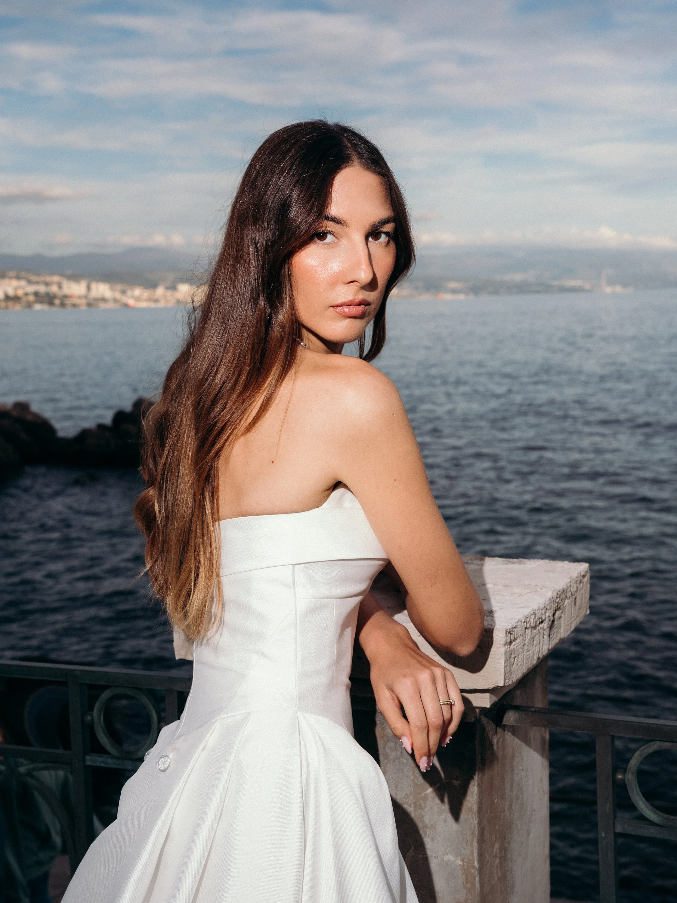 A young woman with long brown hair wearing a white strapless dress, standing by a lake or sea with a city skyline and mountains in the background.