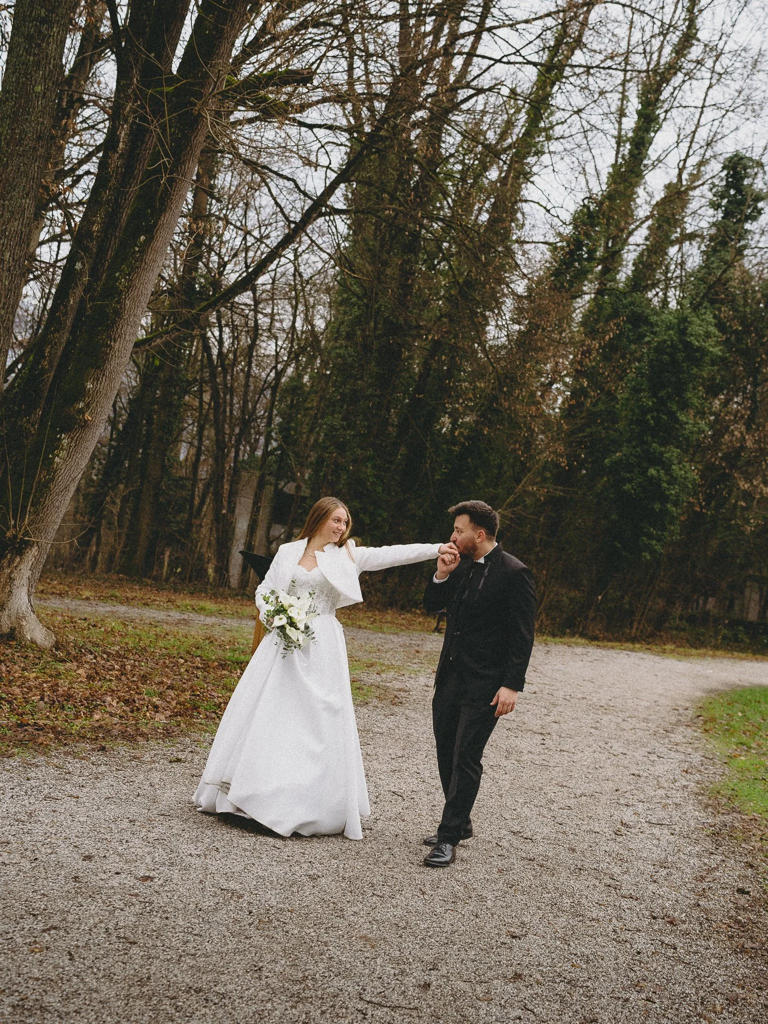 A bride in a white wedding dress and a groom in a black suit sharing a kiss outside on a gravel path surrounded by trees.
