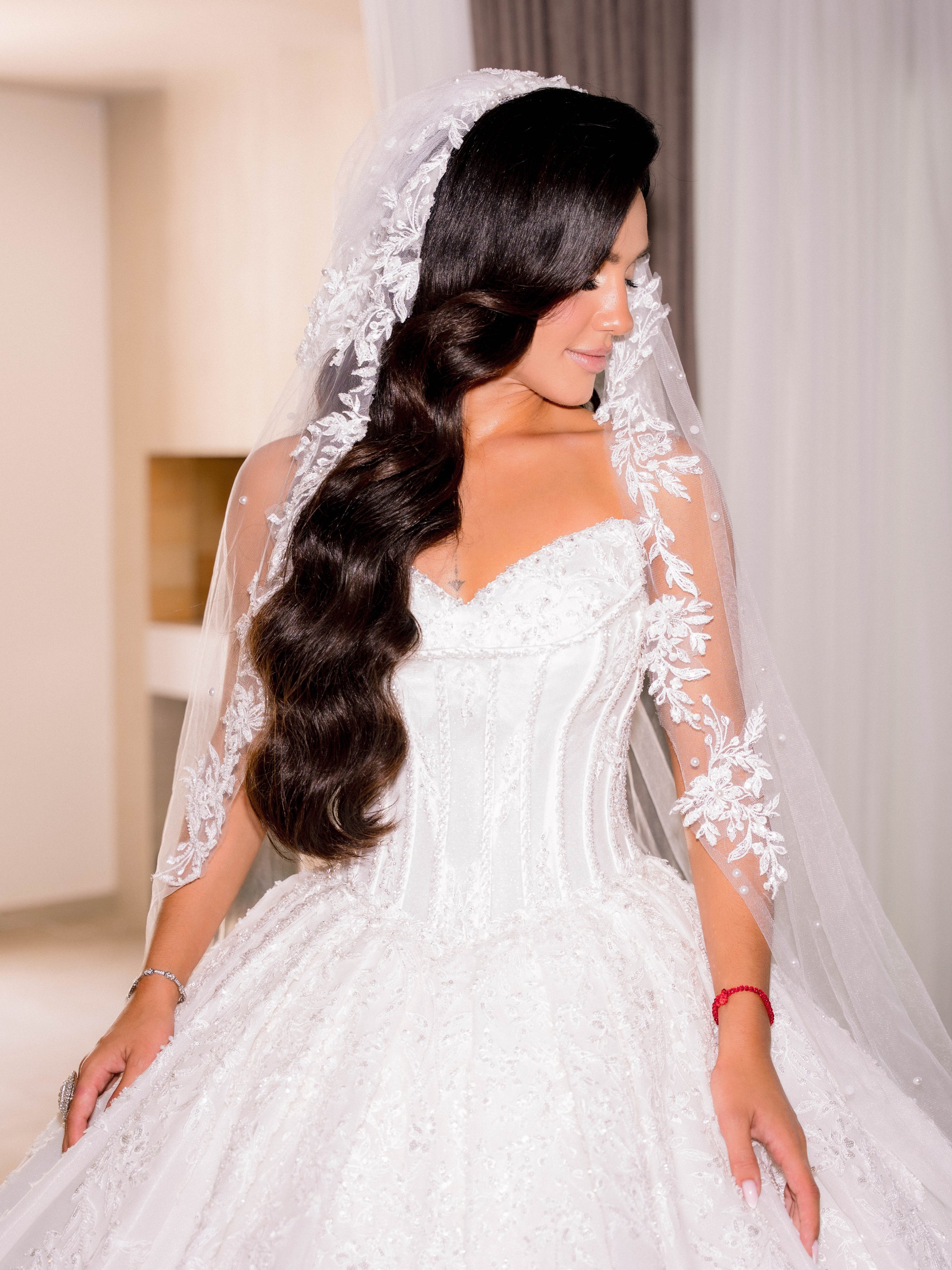 A woman in a white wedding dress with lace details and a sheer veil, with long dark wavy hair, standing indoors with her eyes closed and a serene expression.