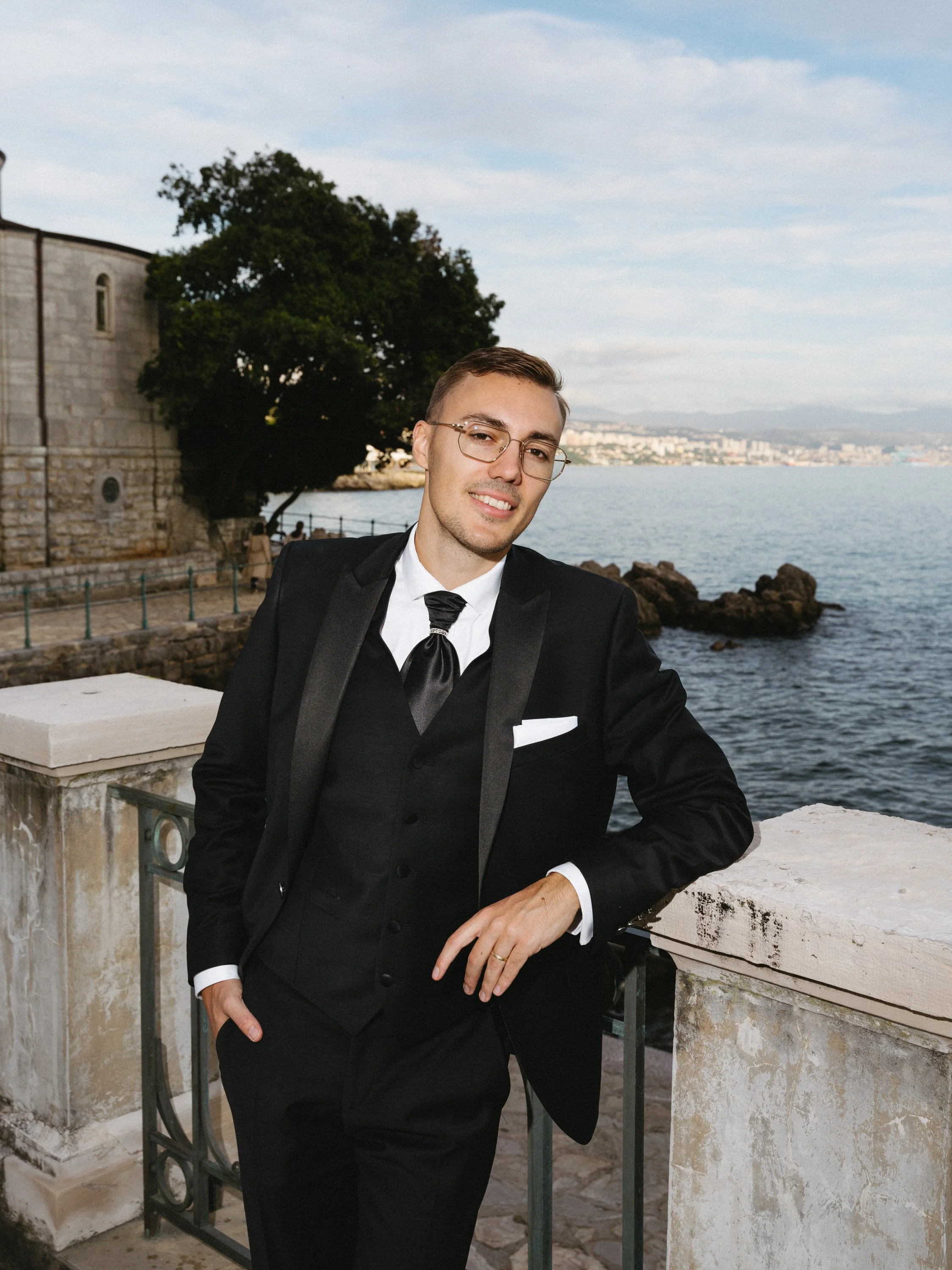 A young man in a black tuxedo with a white shirt and black tie, wearing glasses, smiling slightly while leaning against a stone railing near the water, with a large tree, stone buildings, and a cityscape in the background.