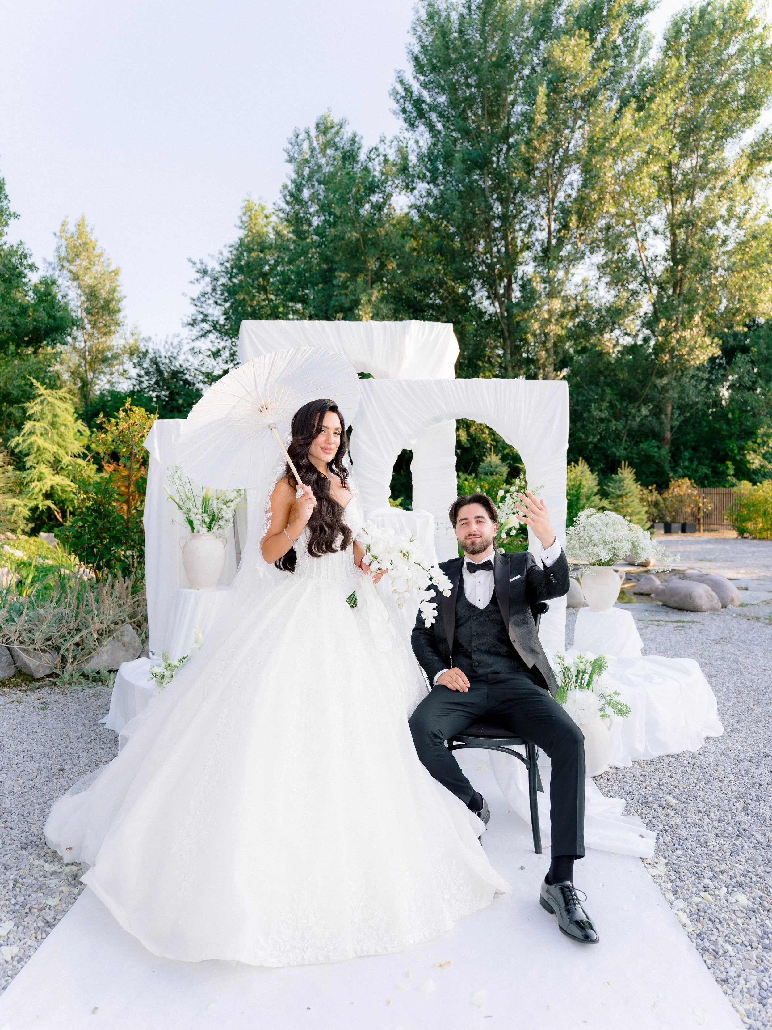 A bride in a wedding dress and a groom in a tuxedo sitting and standing in an outdoor wedding setting with white decorations and potted plants, greenery, and trees in the background.