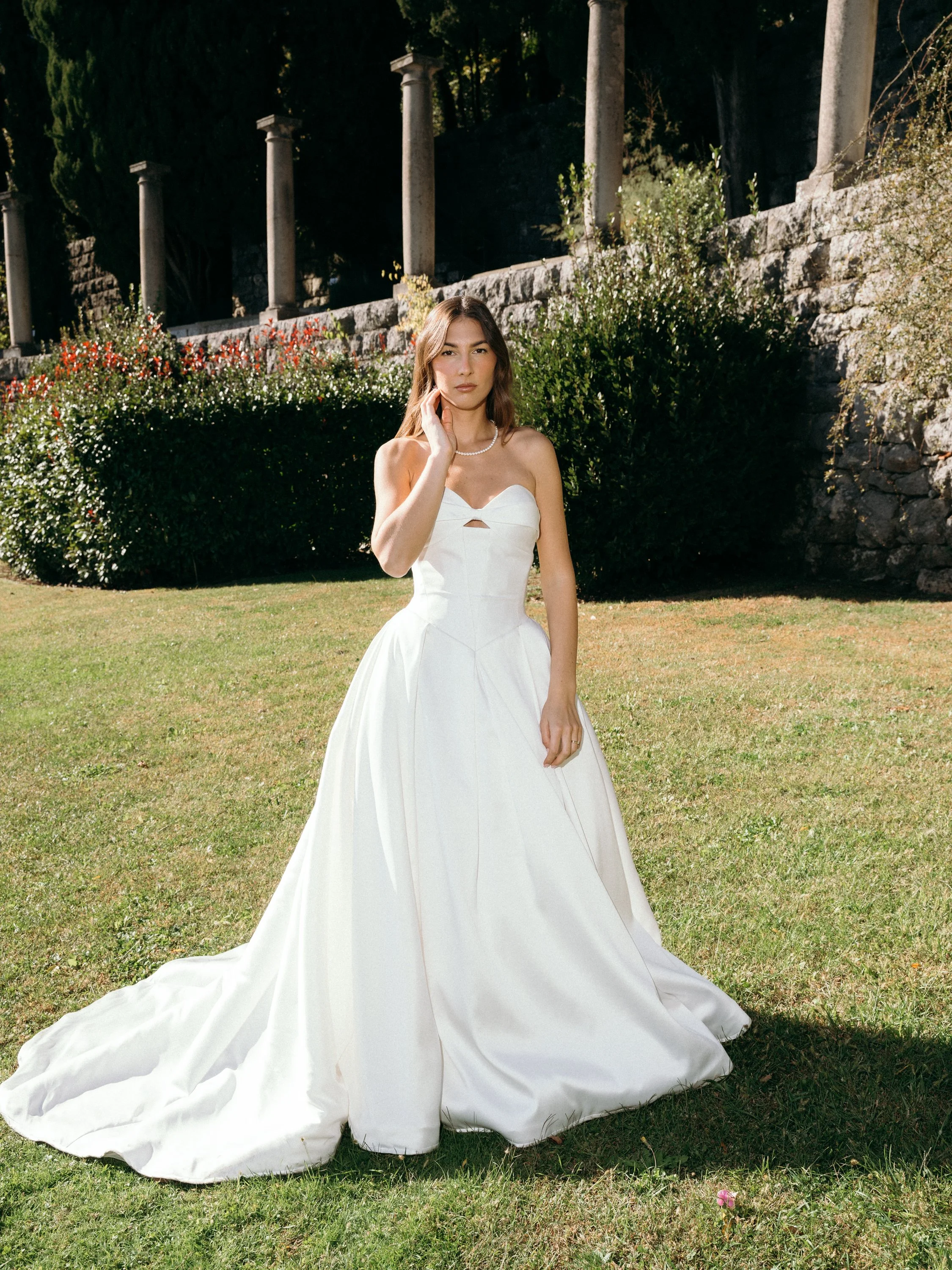 A woman in a white wedding dress standing on a grassy lawn with bushes and stone structures in the background.