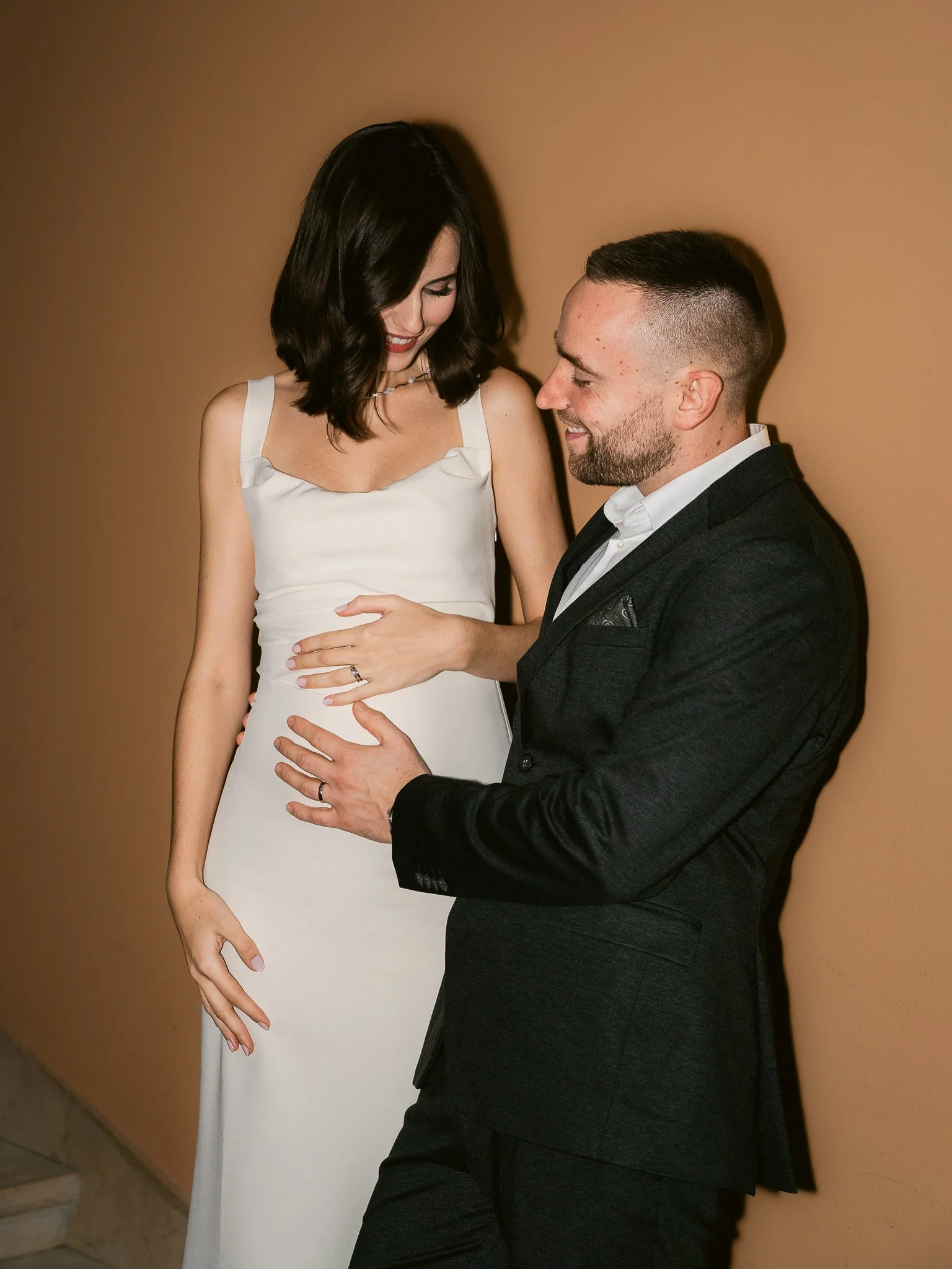 A woman in a white dress and a man in a dark suit are gazing at each other affectionately, with the woman touching the man's chest and the man holding her around the waist. They are smiling, standing against a peach-colored wall.