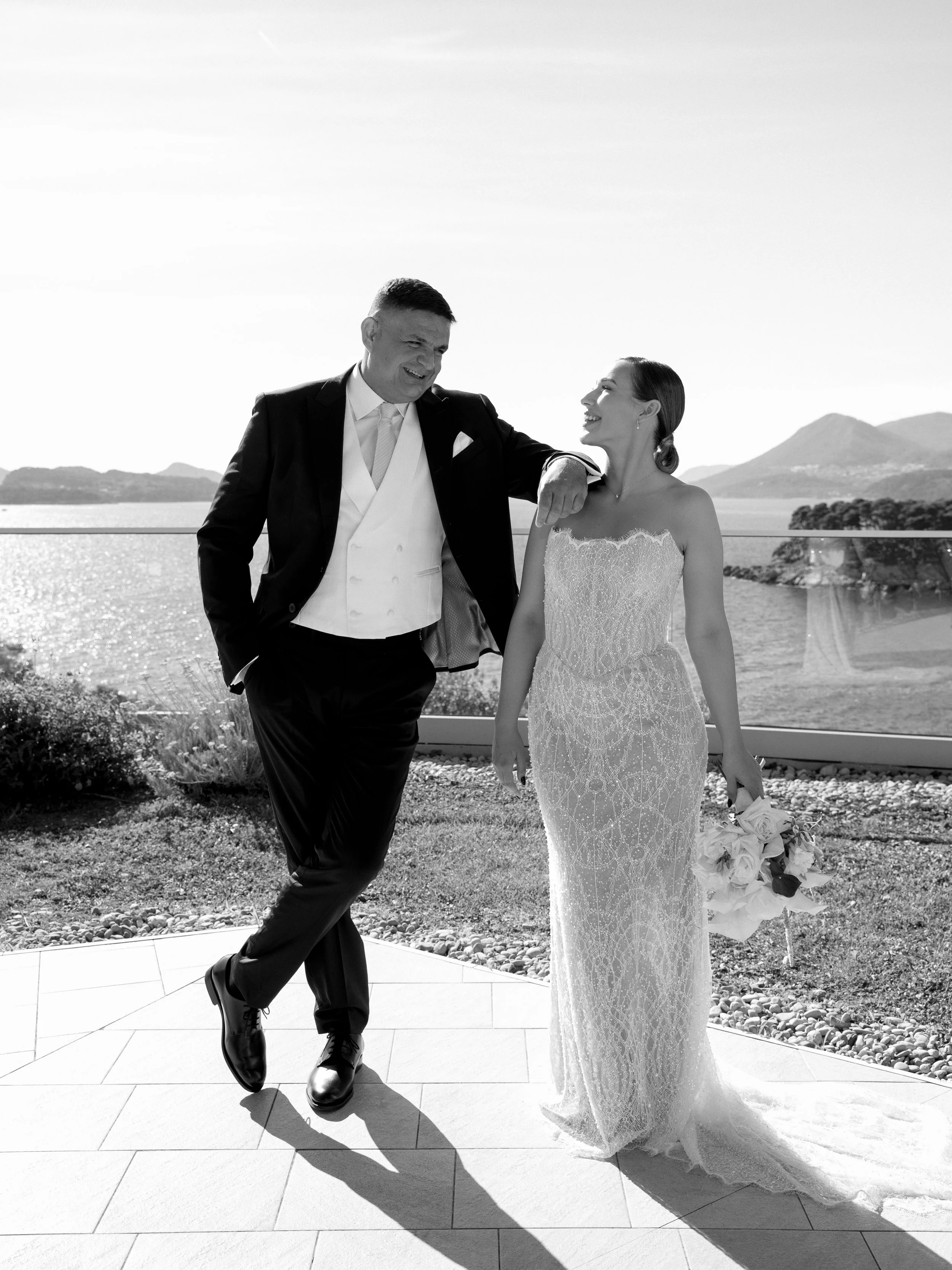 Black and white photo of a bride and groom enjoying a moment together outdoors, with mountains and water in the background.