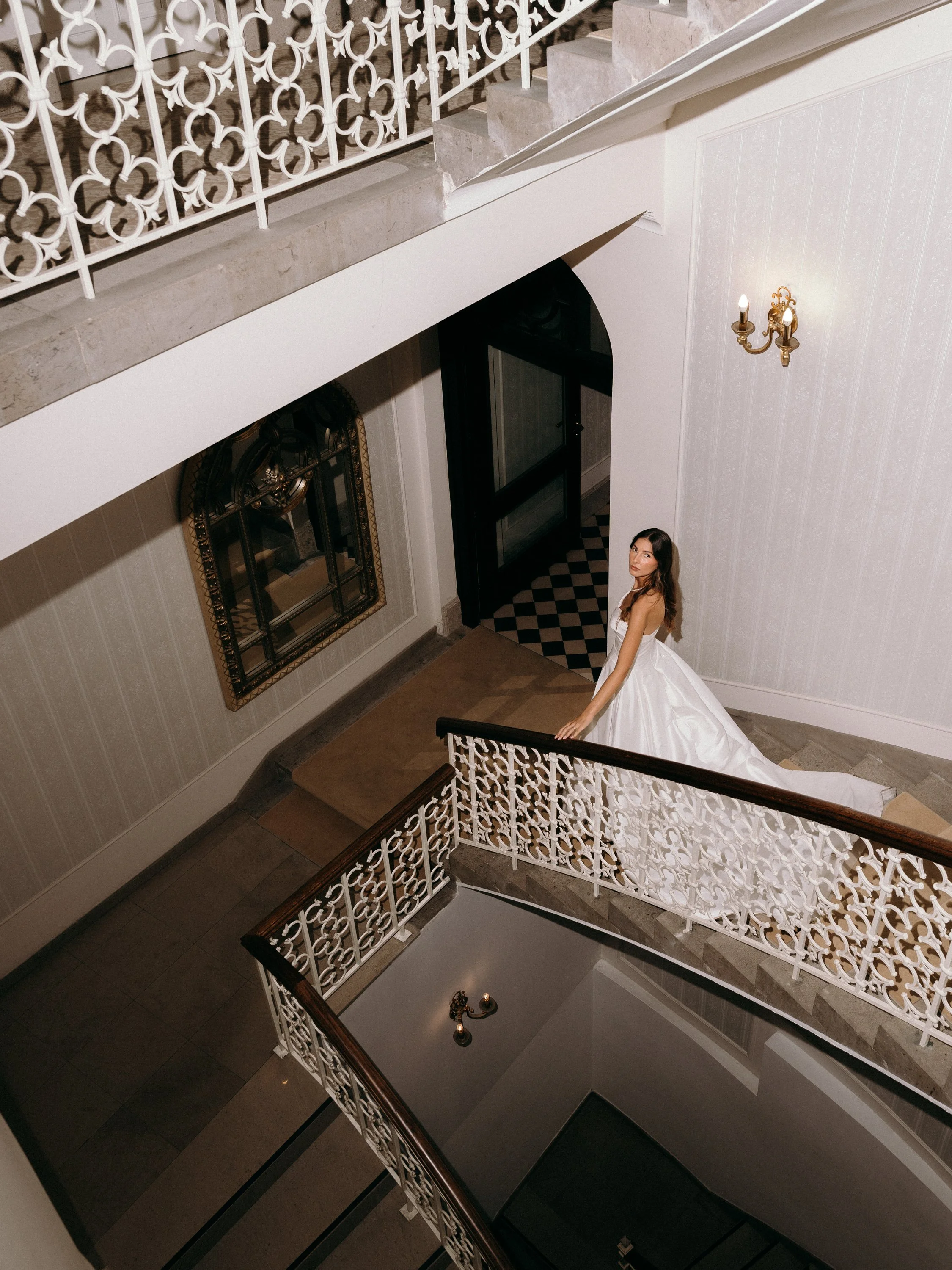 A woman in a white wedding gown standing on a staircase landing inside a building with ornate white railings and vintage decor.