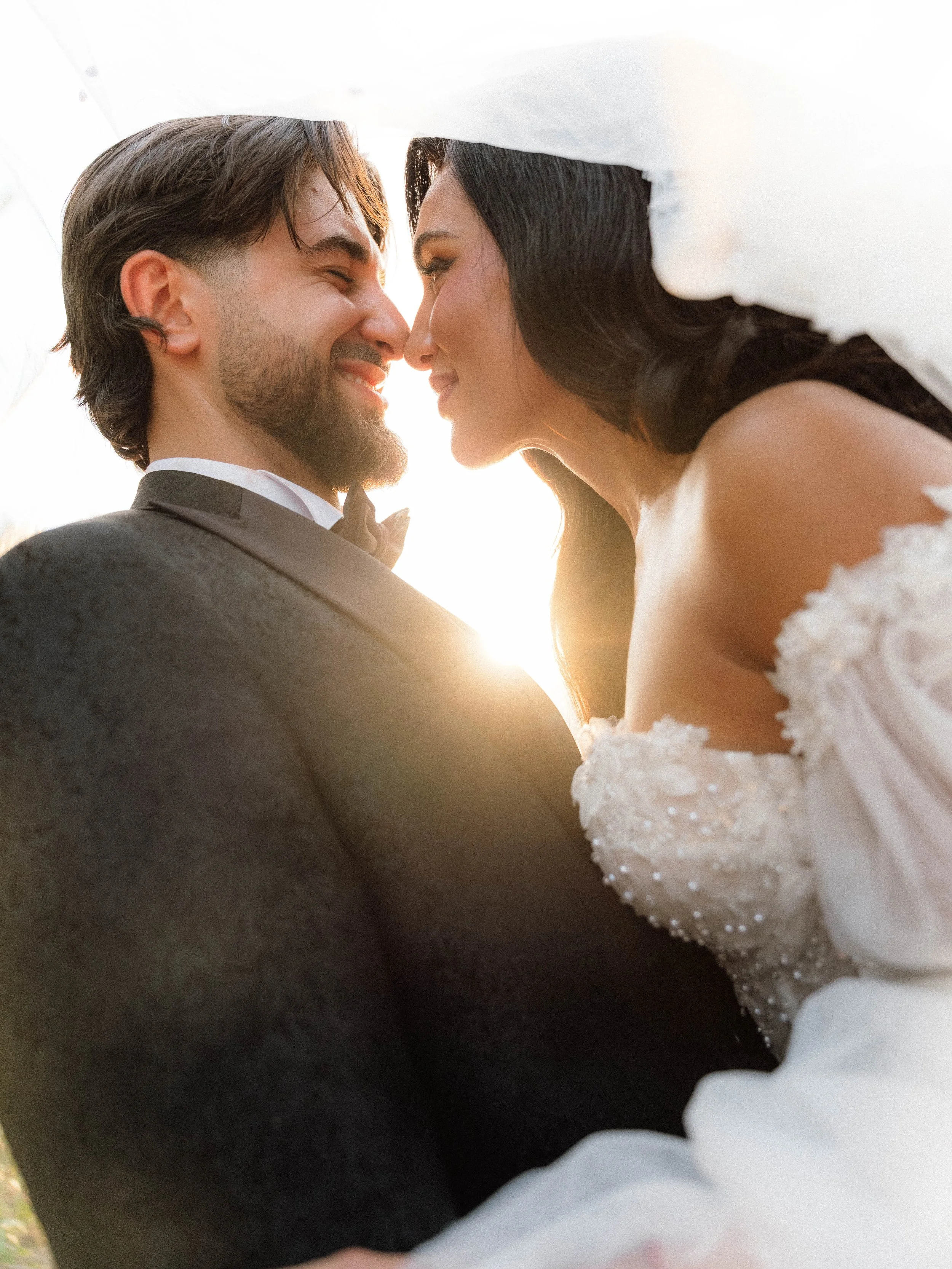 A close-up photograph of a bride and groom gazing into each other's eyes under a white umbrella, with warm sunlight creating a romantic atmosphere.