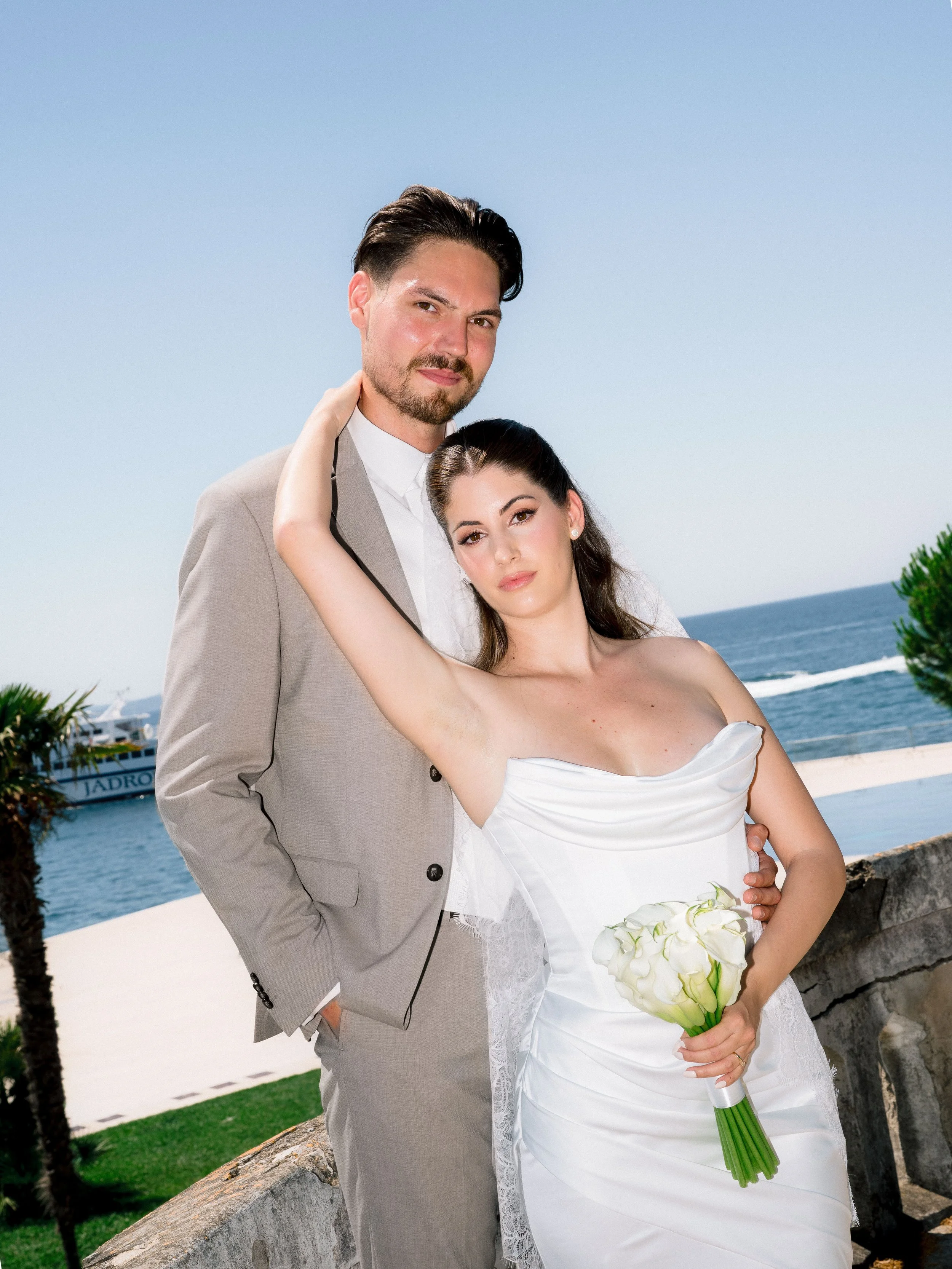 A newlywed couple in wedding attire, standing near the coast, with the ocean in the background. The groom is in a beige suit, and the bride is in a white wedding dress holding a bouquet of white calla lilies.