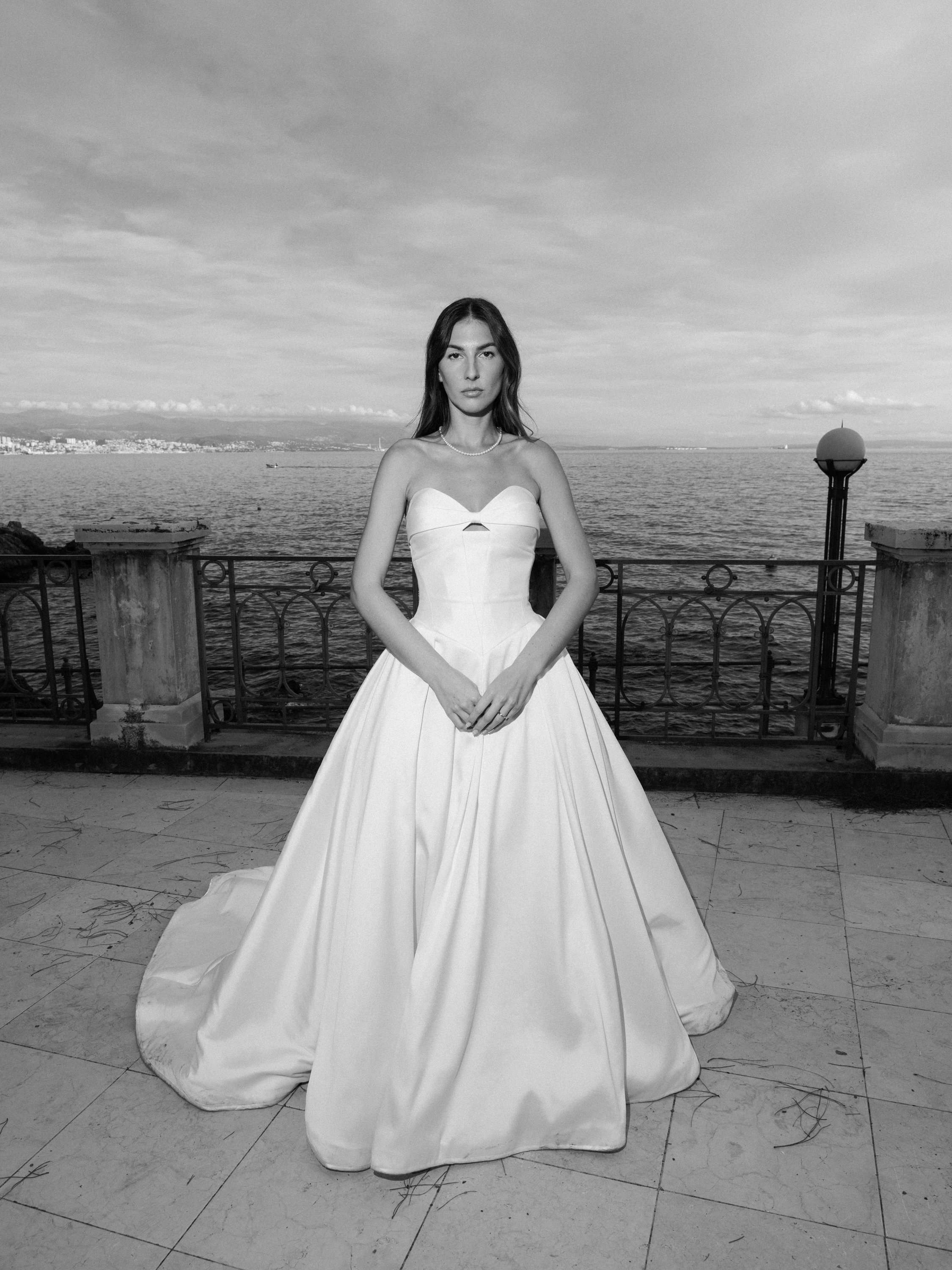 A woman in a wedding dress standing on a terrace with a lake and city skyline in the background, black and white photo.