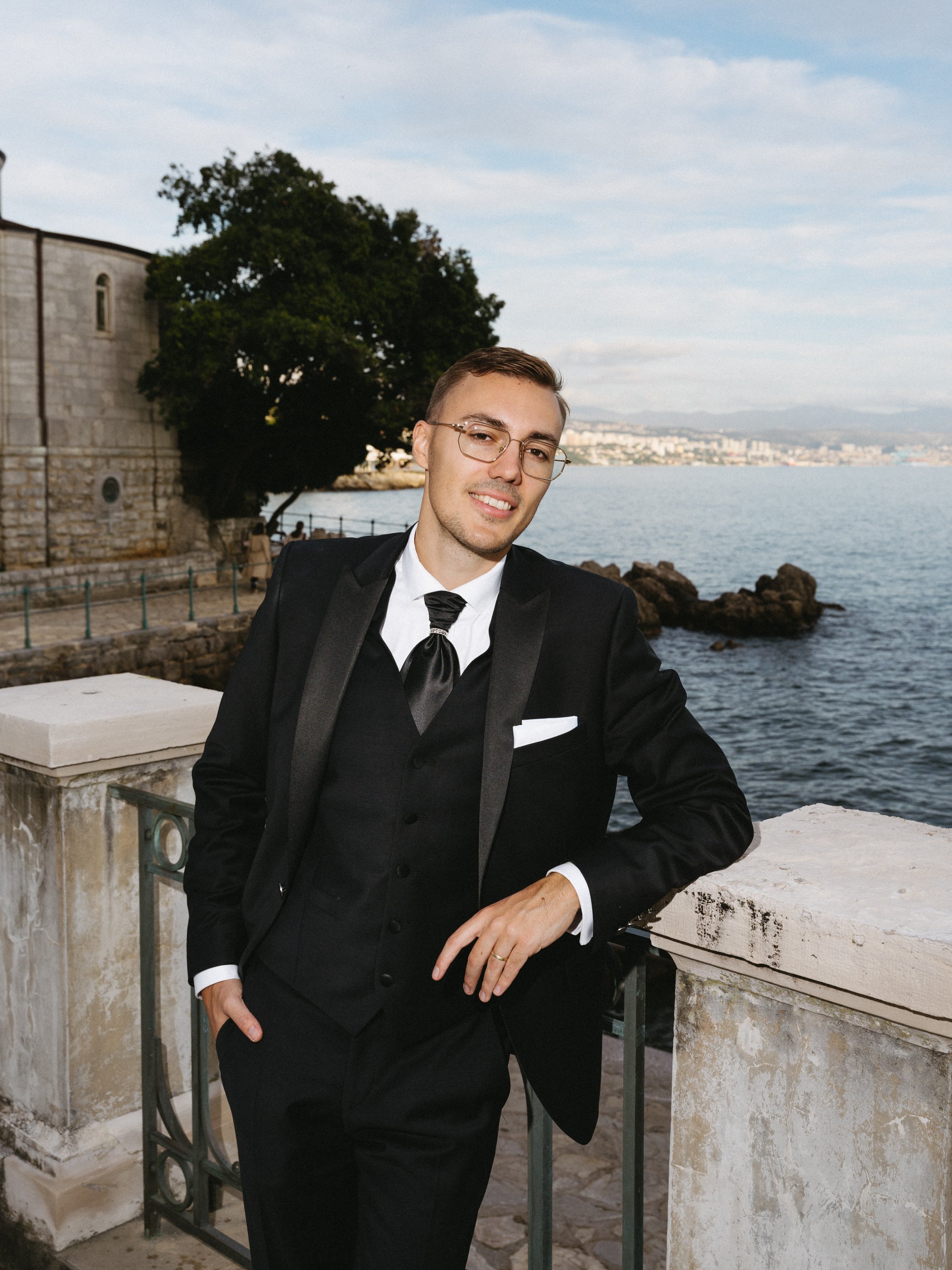 A young man in a black tuxedo with a white shirt and black tie standing by a stone railing near the water, with a cityscape and mountains in the background.