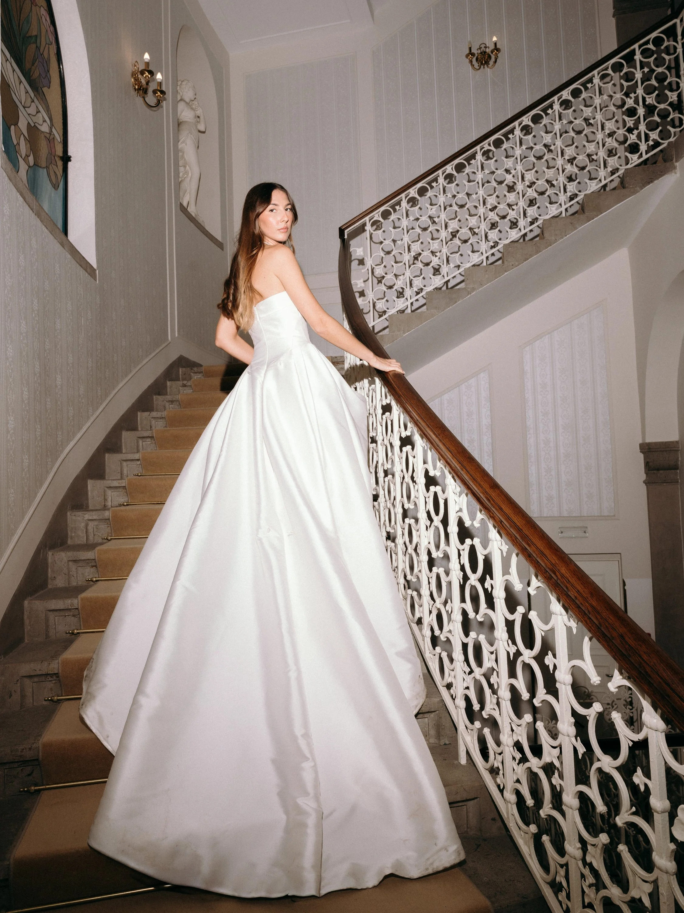A woman in a strapless white gown standing on a staircase with ornate white railing, with wall lamps and statues in the background.