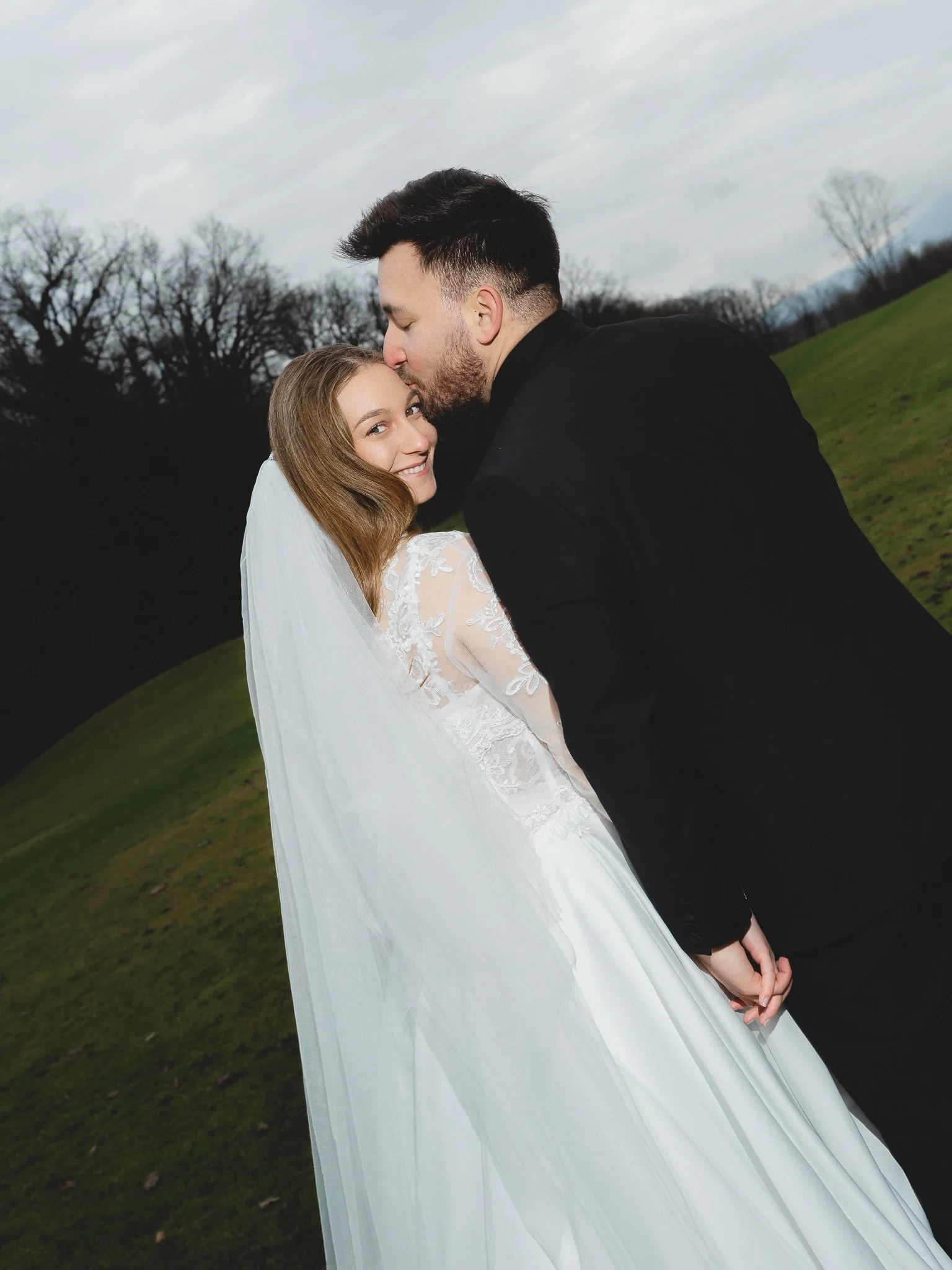 A bride and groom holding hands outdoors, with the groom kissing the bride on the forehead, against a backdrop of trees and a cloudy sky.