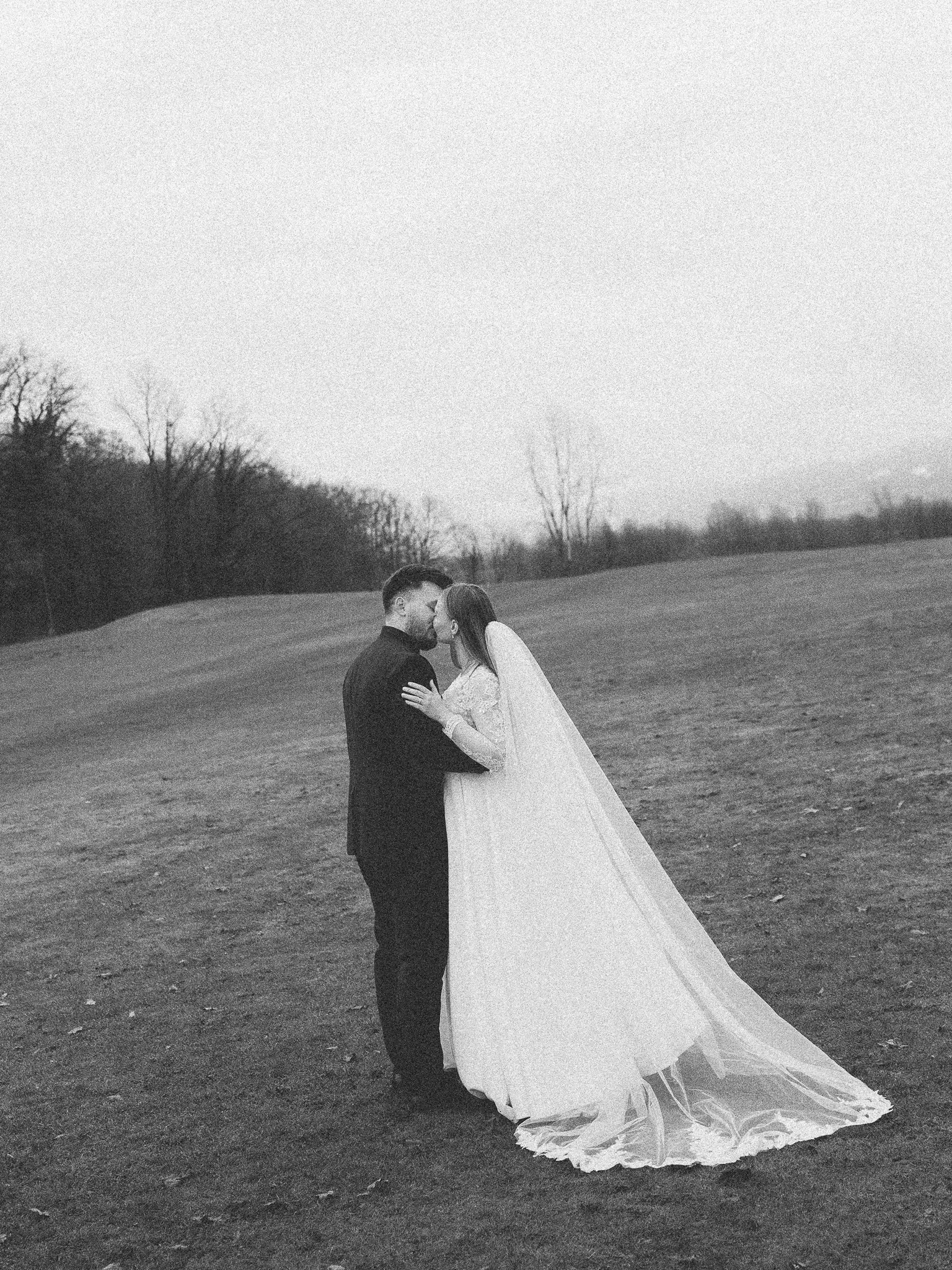A black and white photograph of a newlywed couple standing and embracing outdoors in an open field with trees in the background, sharing a kiss.