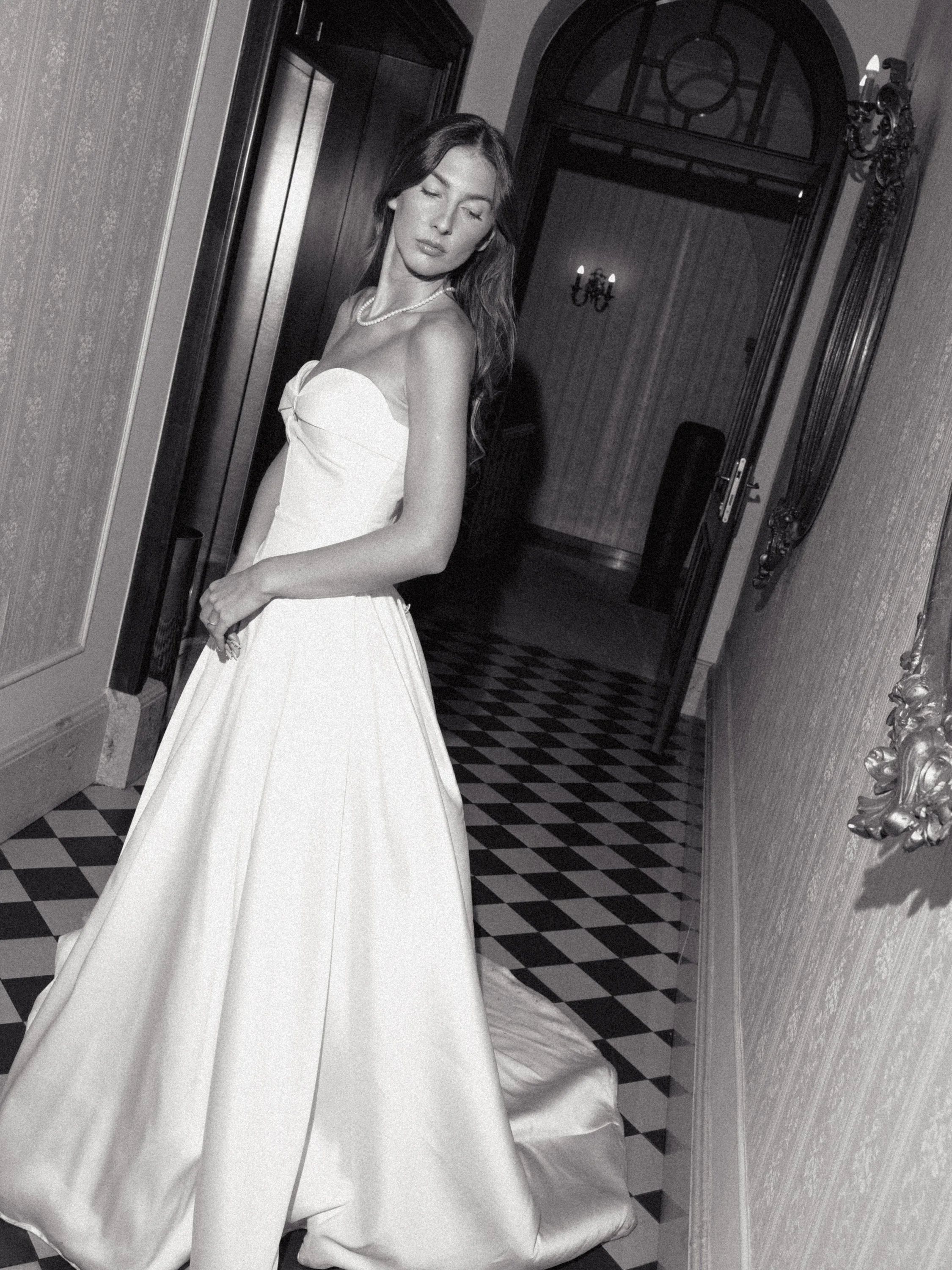 A woman in a strapless white gown with a full skirt stands in a vintage hallway, looking down with her eyes closed. She wears a pearl necklace, and her long hair cascades over her shoulders. The hallway features checkered flooring, ornate wallpaper, 