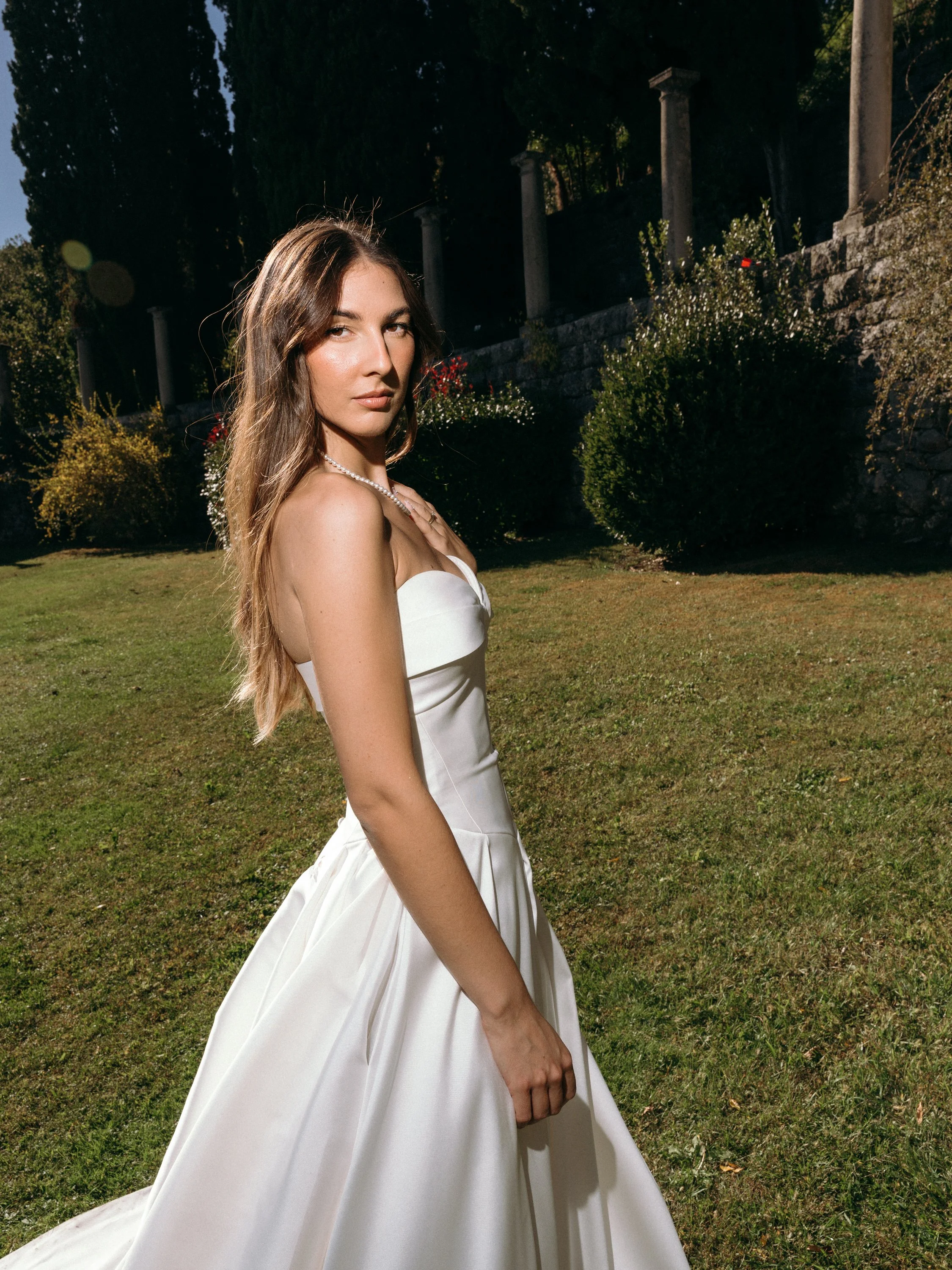 A young woman in a white wedding dress stands outdoors on a grassy lawn with trees, shrubs, and stone pillars in the background.