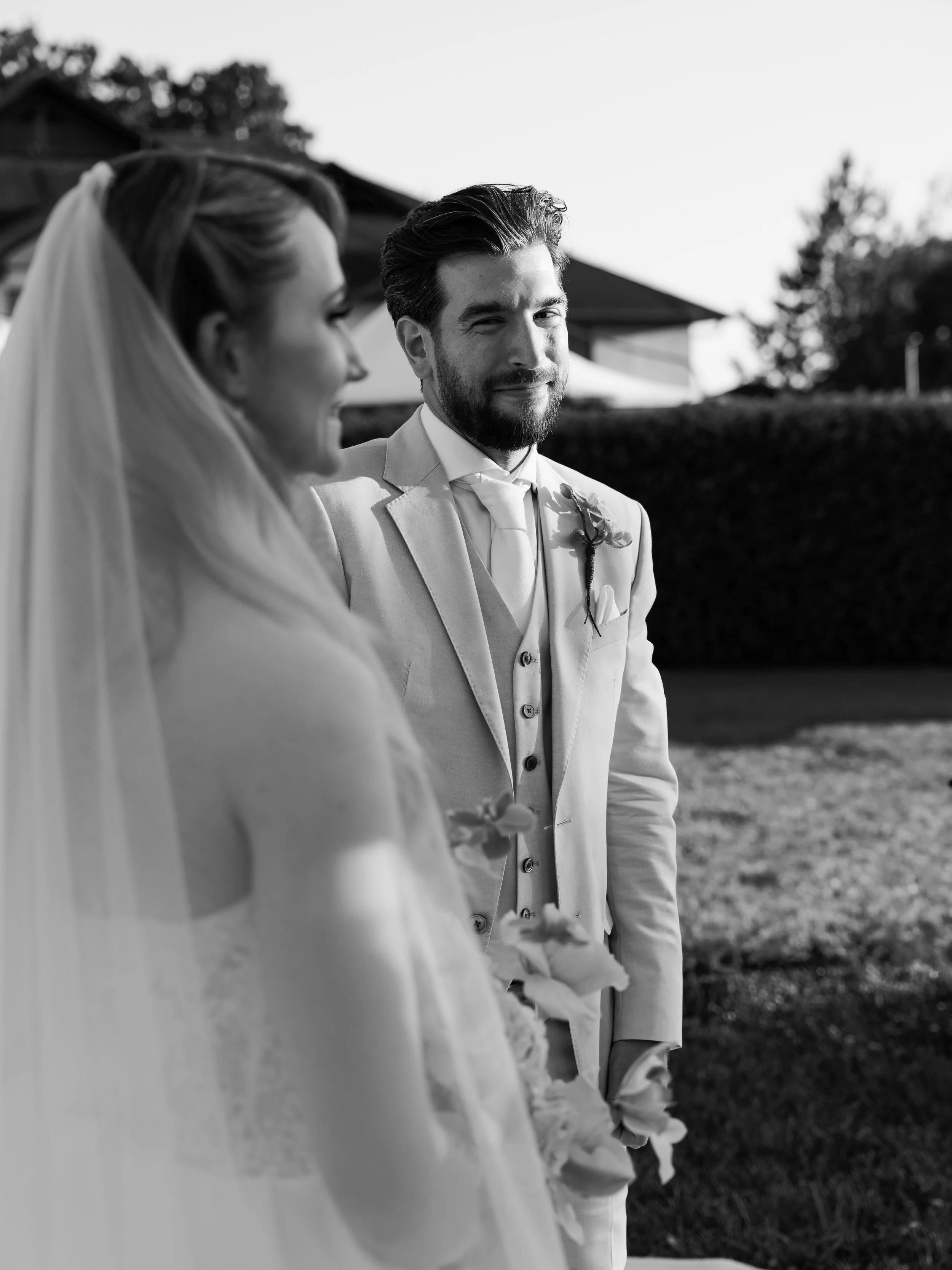 Black and white photo of a bride and groom outdoors, with the groom winking and smiling, in front of a building and trees.