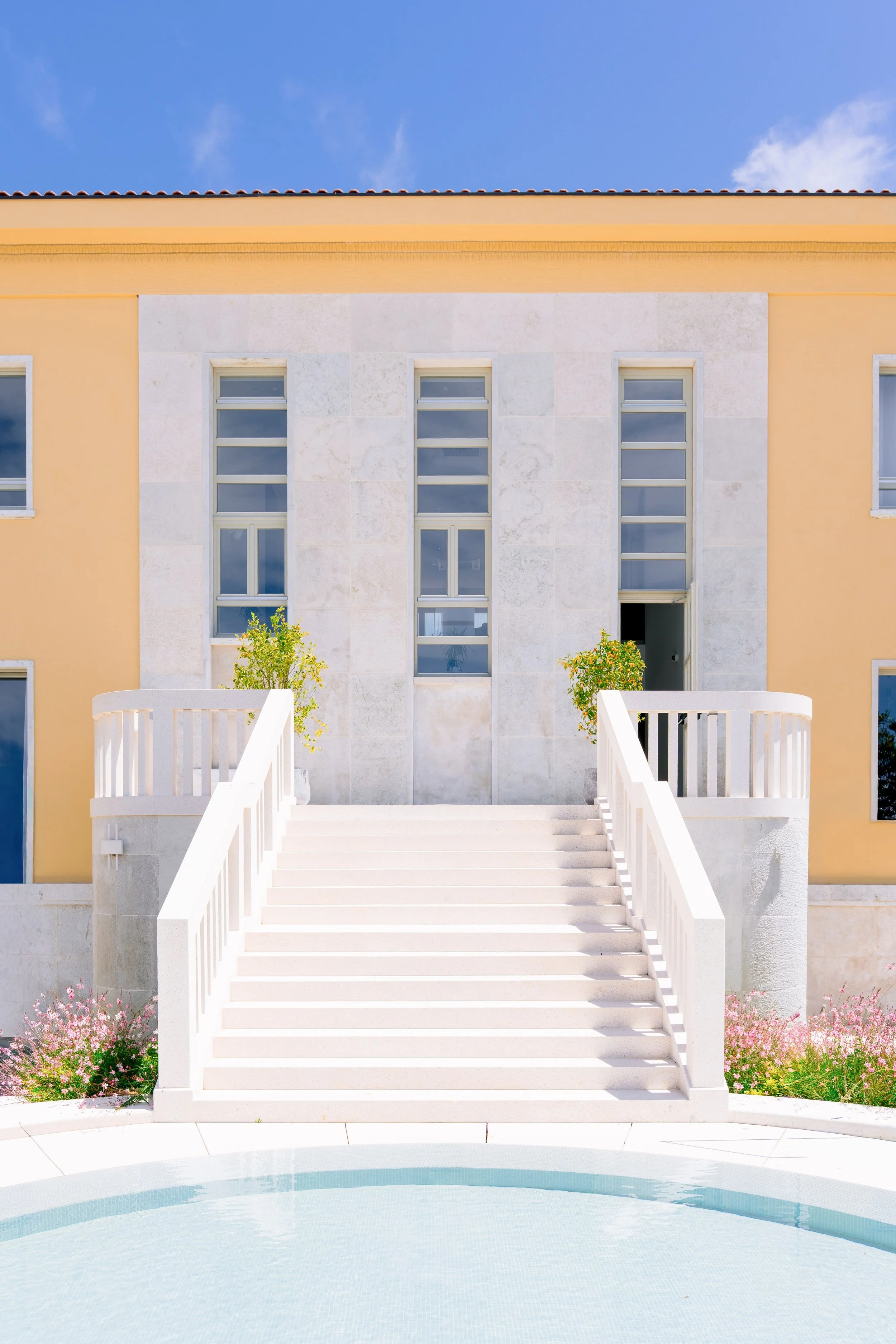 Front view of a yellow and white house with a staircase leading to the entrance, flanked by small trees and pink flowers, with a swimming pool at the front and a blue sky with a few clouds above.
