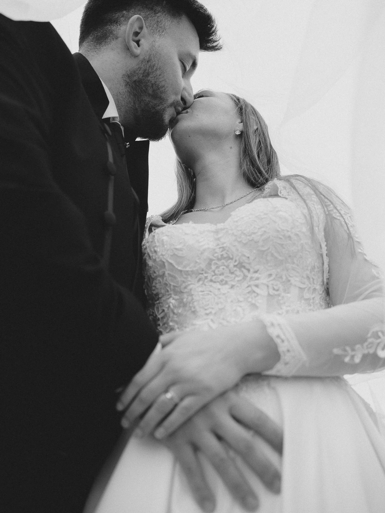 Black and white photo of a couple kissing, with the man wearing a dark suit and the woman wearing a lace dress, with her hand resting on his arm.
