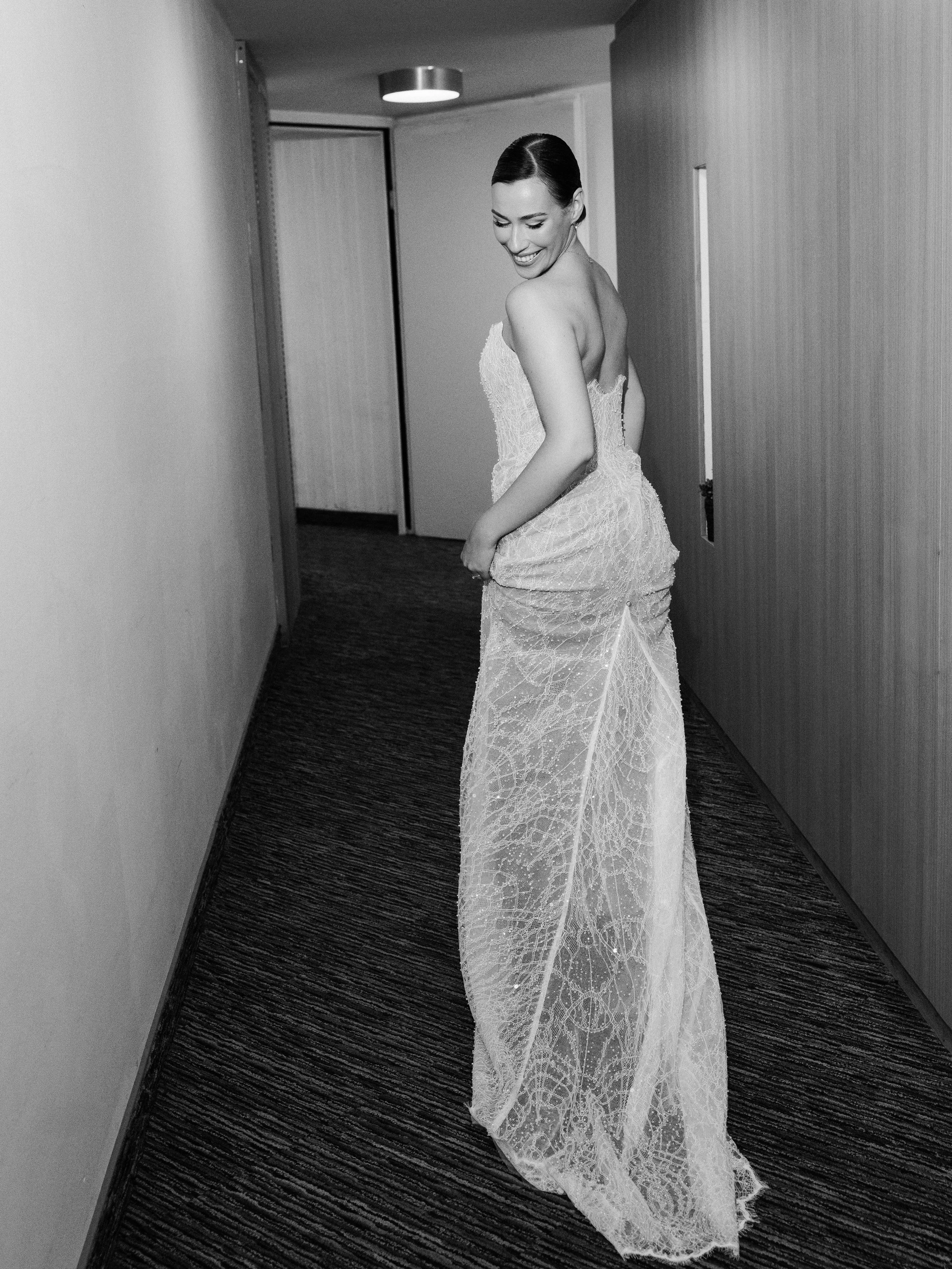 A woman in a wedding dress standing in a hallway, smiling and looking to her left.