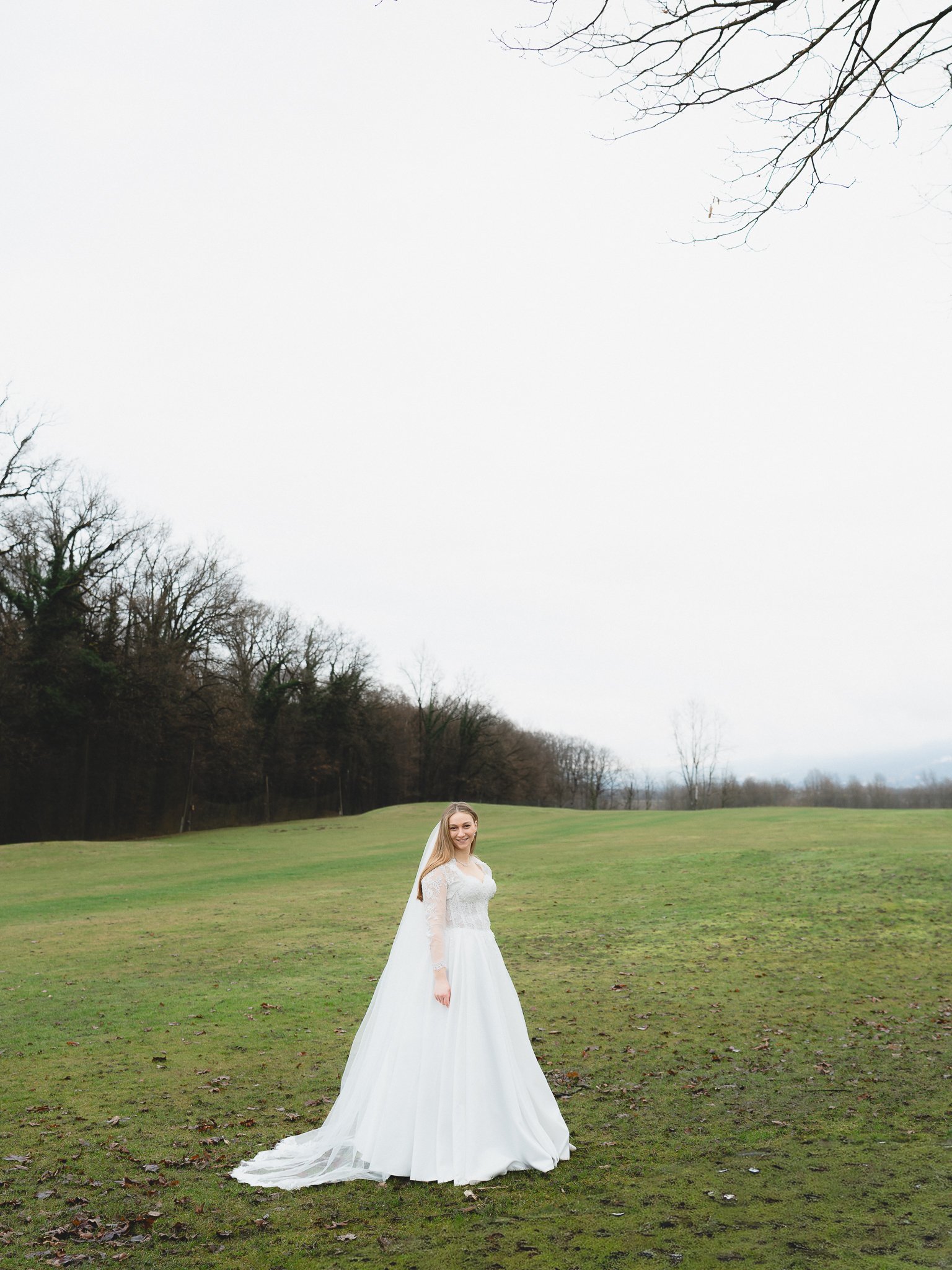 A bride in a white wedding dress standing in an open grassy field with trees and overcast sky in the background.