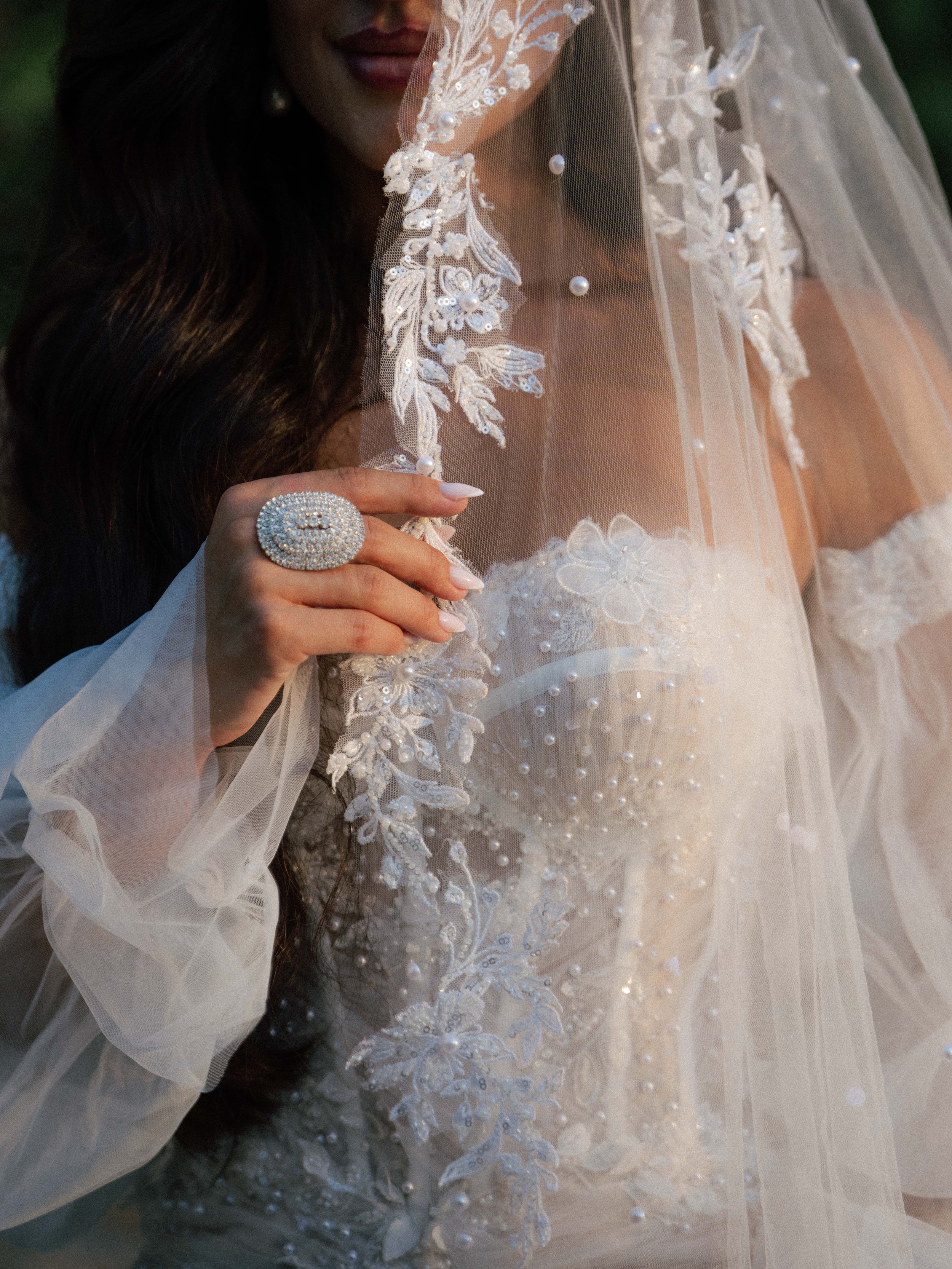 Close-up of a woman dressed in a wedding gown with detailed lace and bead embroidery, wearing a sheer veil with pearl accents, and a large ring with a cluster of pearls and crystals.