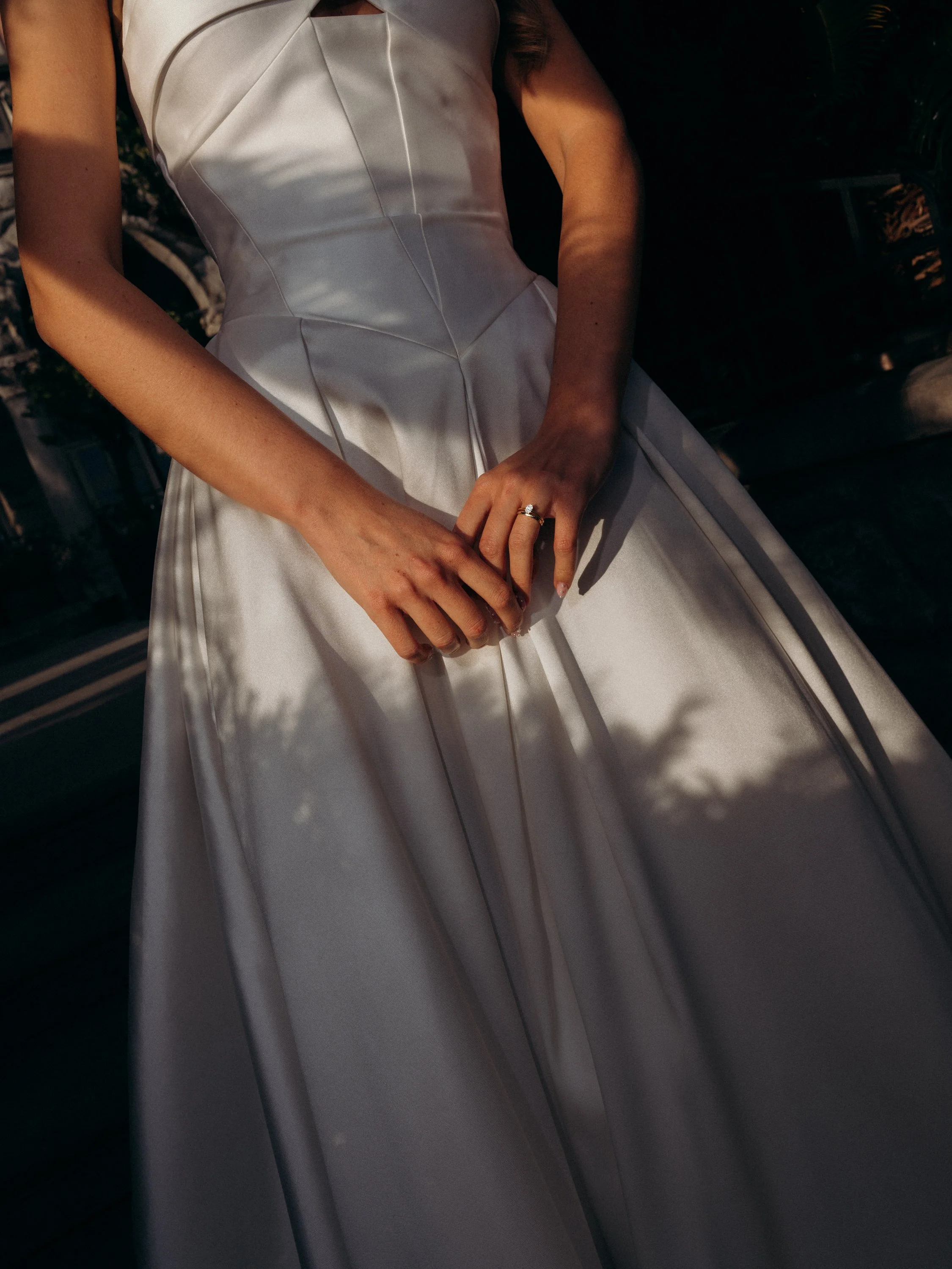 A woman in a white gown with detailed seams, sitting outdoors with shadows of trees cast on her dress, displaying a wedding ring on her left hand.