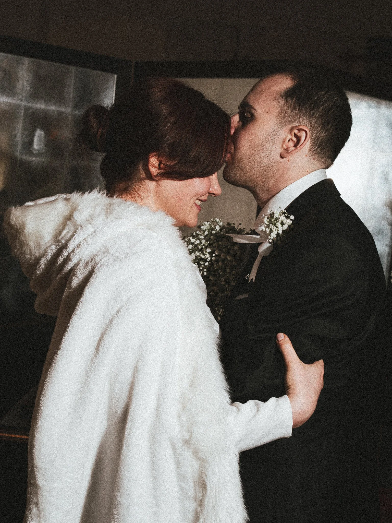 A couple sharing a tender moment, with eyes closed and foreheads touching, one wearing a white fur coat and the other in a black suit with boutonniere, inside a dimly lit room.