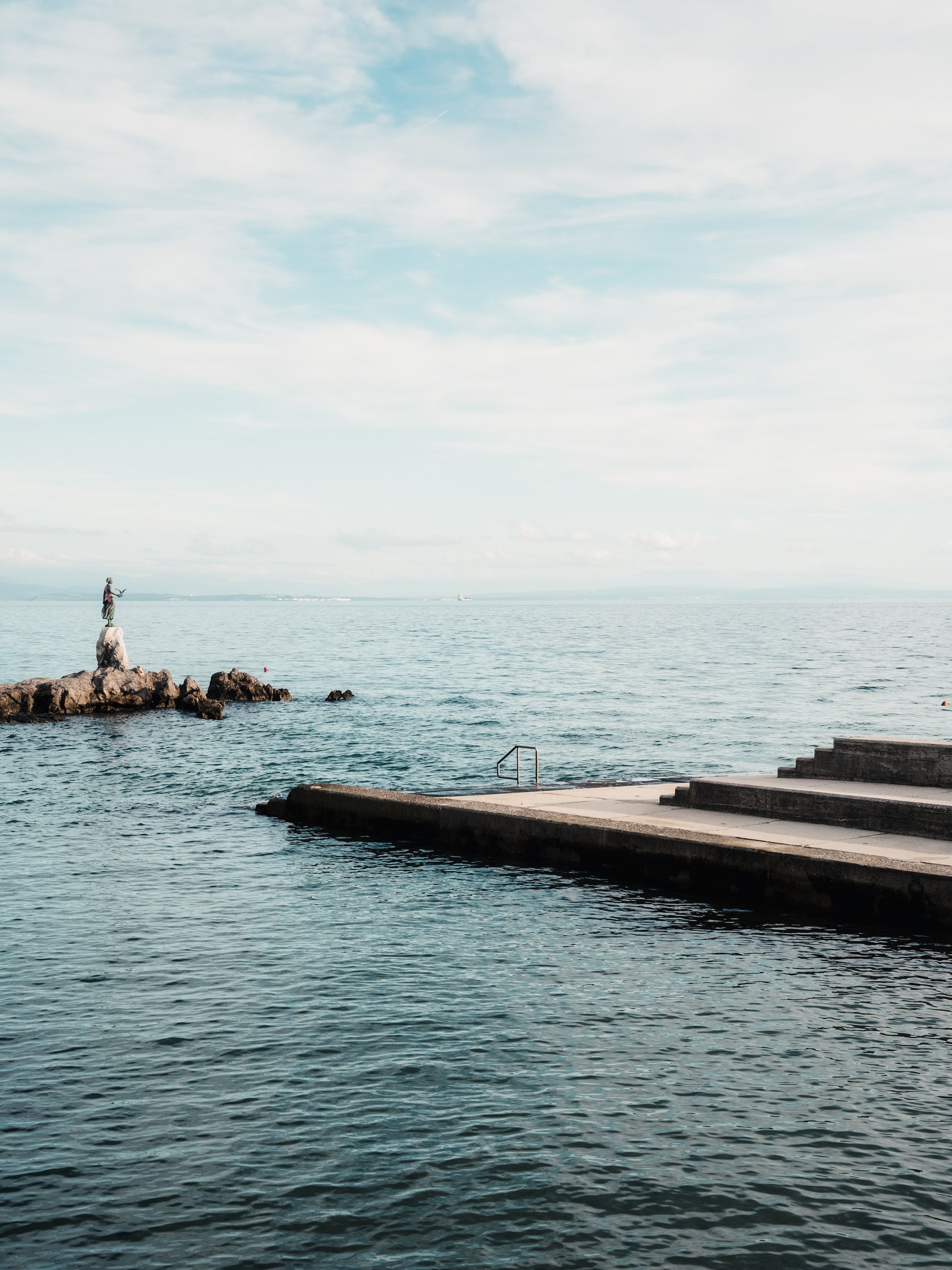 Calm sea with a concrete dock and stairs, and a statue of a woman on a rock formation in the water under a cloudy sky.