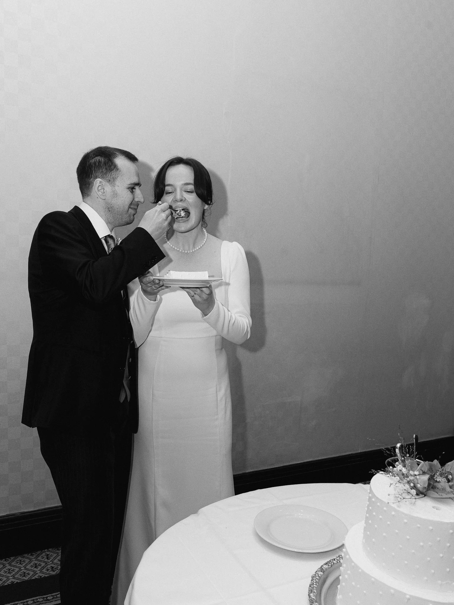 A man in a suit feeds a slice of cake to a woman in a white dress at a celebration, with a cake on the table nearby.