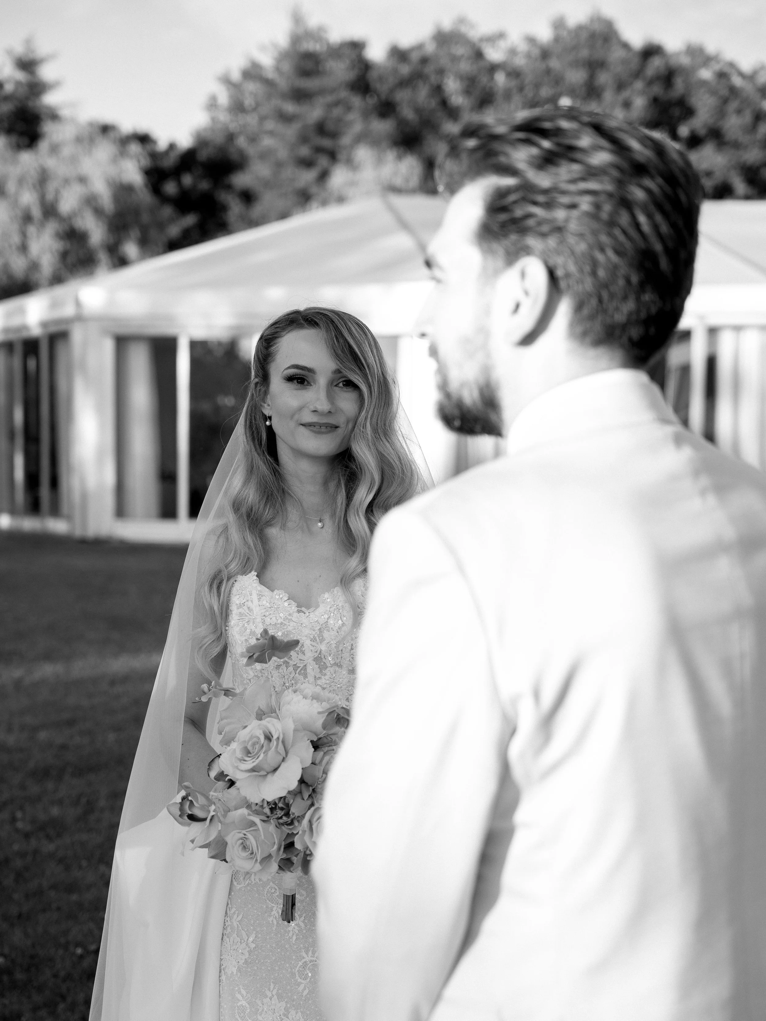 A bride holding a bouquet of flowers looks at a groom. The photo is in black and white, taken outdoors with trees and a building in the background.