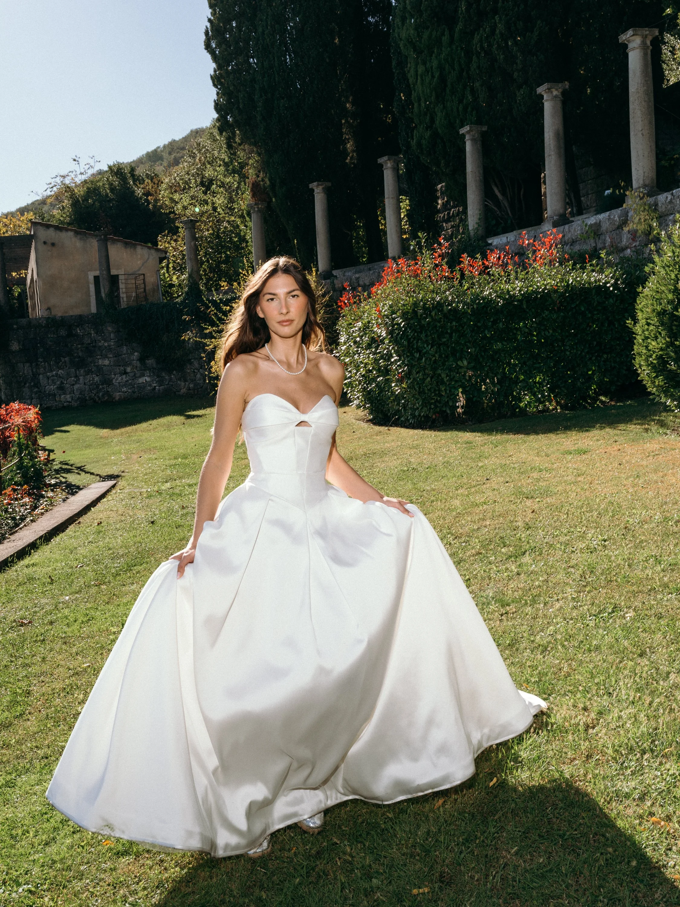 A woman in a white wedding dress standing outdoors on a grassy lawn with trees and columns in the background.
